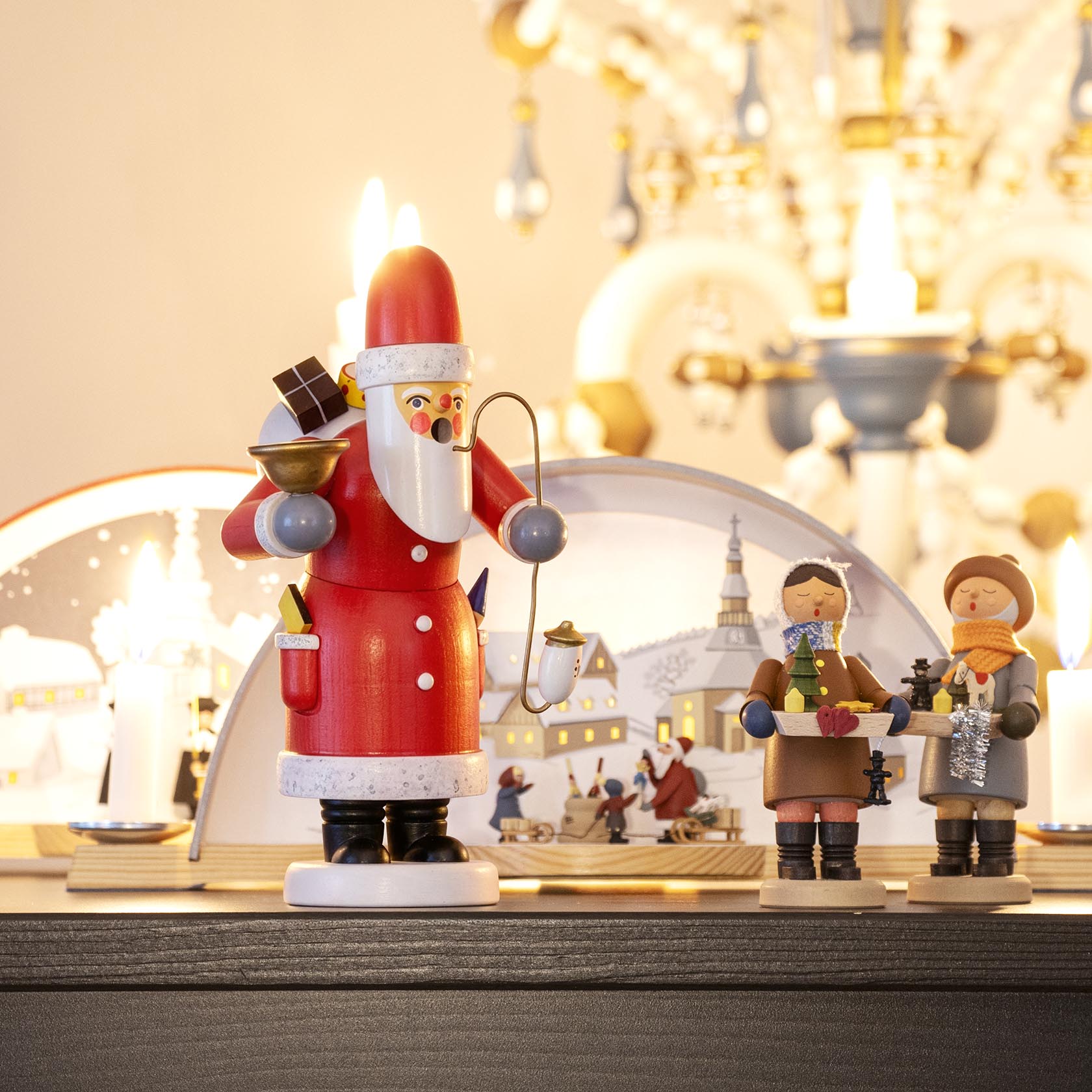 Ein Räuchermann in Form eines Weihnachtsmanns hält eine Pfeife und einen Kerzenhalter. Eine Kerze brennt darauf, daneben stehen zwei weitere Figuren.