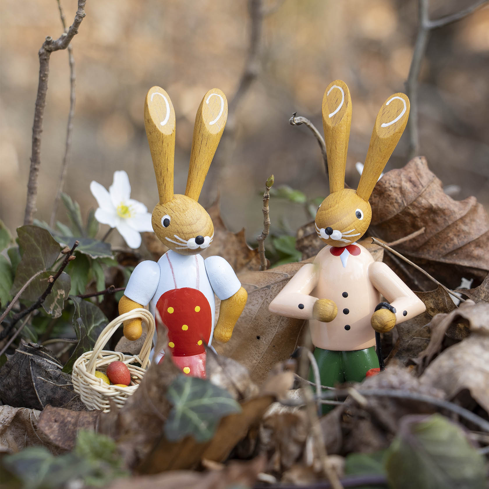 Zwei Holzhasenfiguren im Wald. Der linke Hase trägt eine Schürze und hält einen geflochtenen Korb mit roten Eiern. Der rechte Hase trägt eine Fliege und einen Anzug.