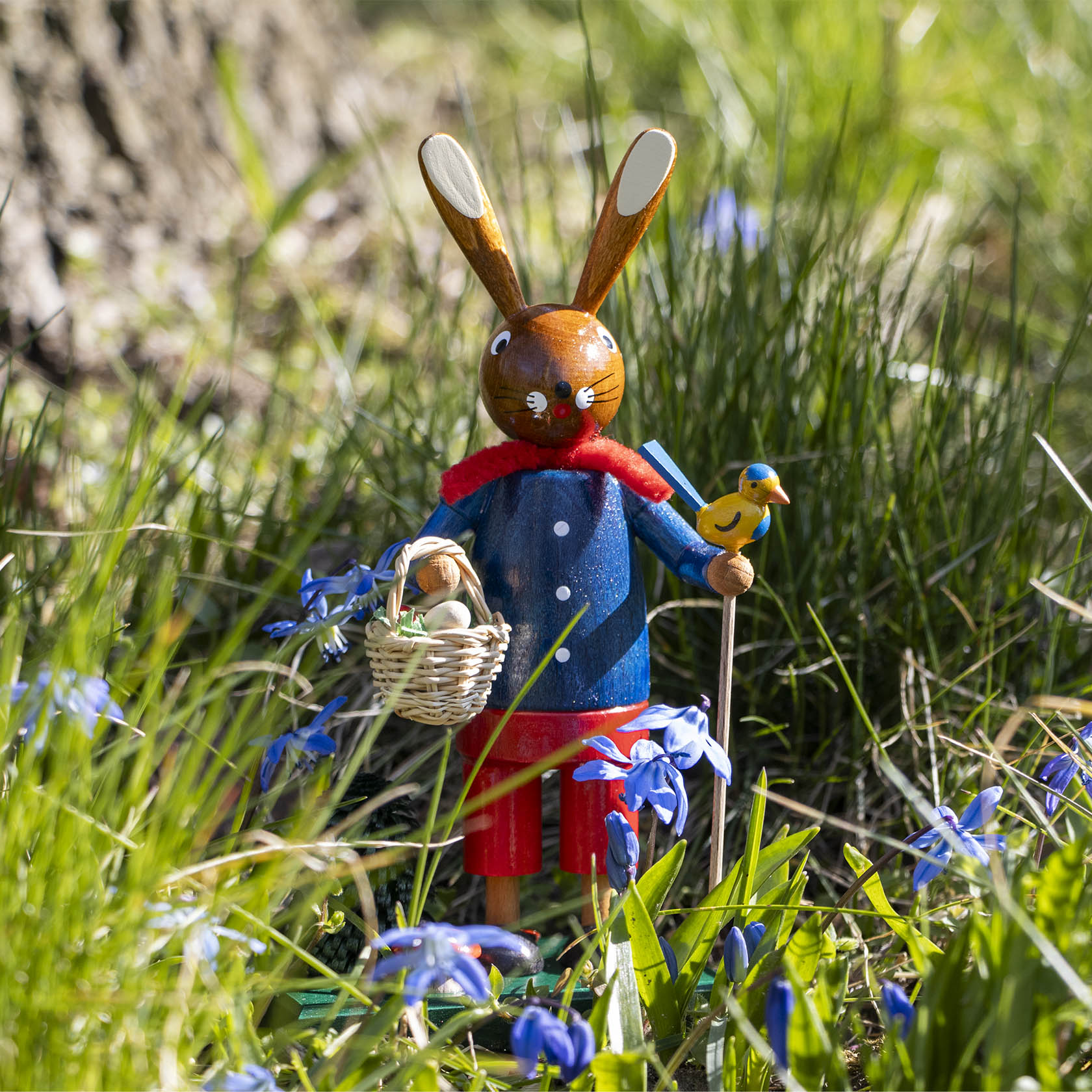 Holzfigur eines Osterhasen in blauem Mantel mit rotem Schal, der einen Korb mit Eiern trägt. Ein kleiner gelber Vogel sitzt auf seiner Armlehne.
