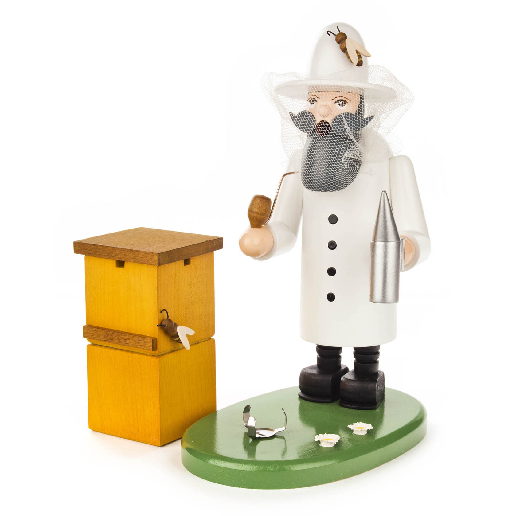 Holzfigur eines Imkers mit schwarzem Bart, weißem Schutzanzug und Schleier. Er hält ein Rauchgerät und steht neben einem Bienenstock auf einem grünen Sockel.