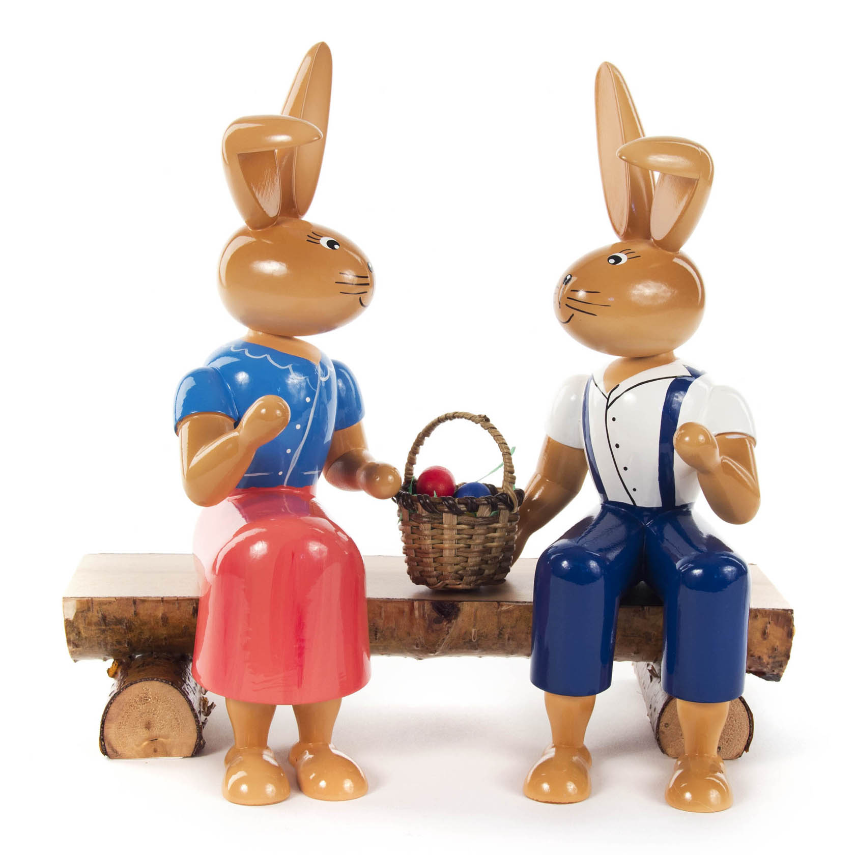 Zwei hölzerne Osterhasen auf einer Holzbank. Der weibliche Hase trägt ein blaues Oberteil und roten Rock, der männliche eine blaue Hose. Ein Korb mit Eiern steht zwischen ihnen. im Dregeno Online Shop günstig kaufen