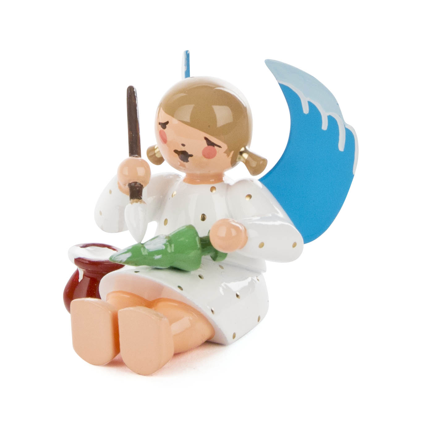 Eine Figur eines sitzenden Engels mit blauem Flügel, der einen Pinsel hält und einen kleinen Weihnachtsbaum grün anmalt. im Dregeno Online Shop günstig kaufen
