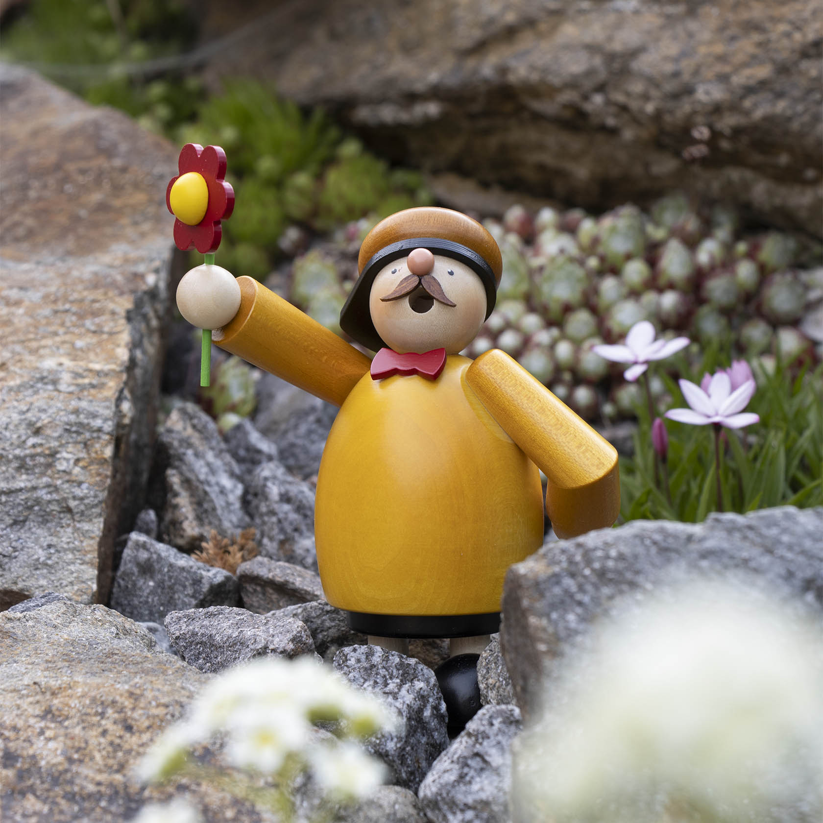A wooden incense smoker figure of a cheerful man with a yellow coat, holding a red flower, standing on rocks. He has a mustache and is wearing a hat.