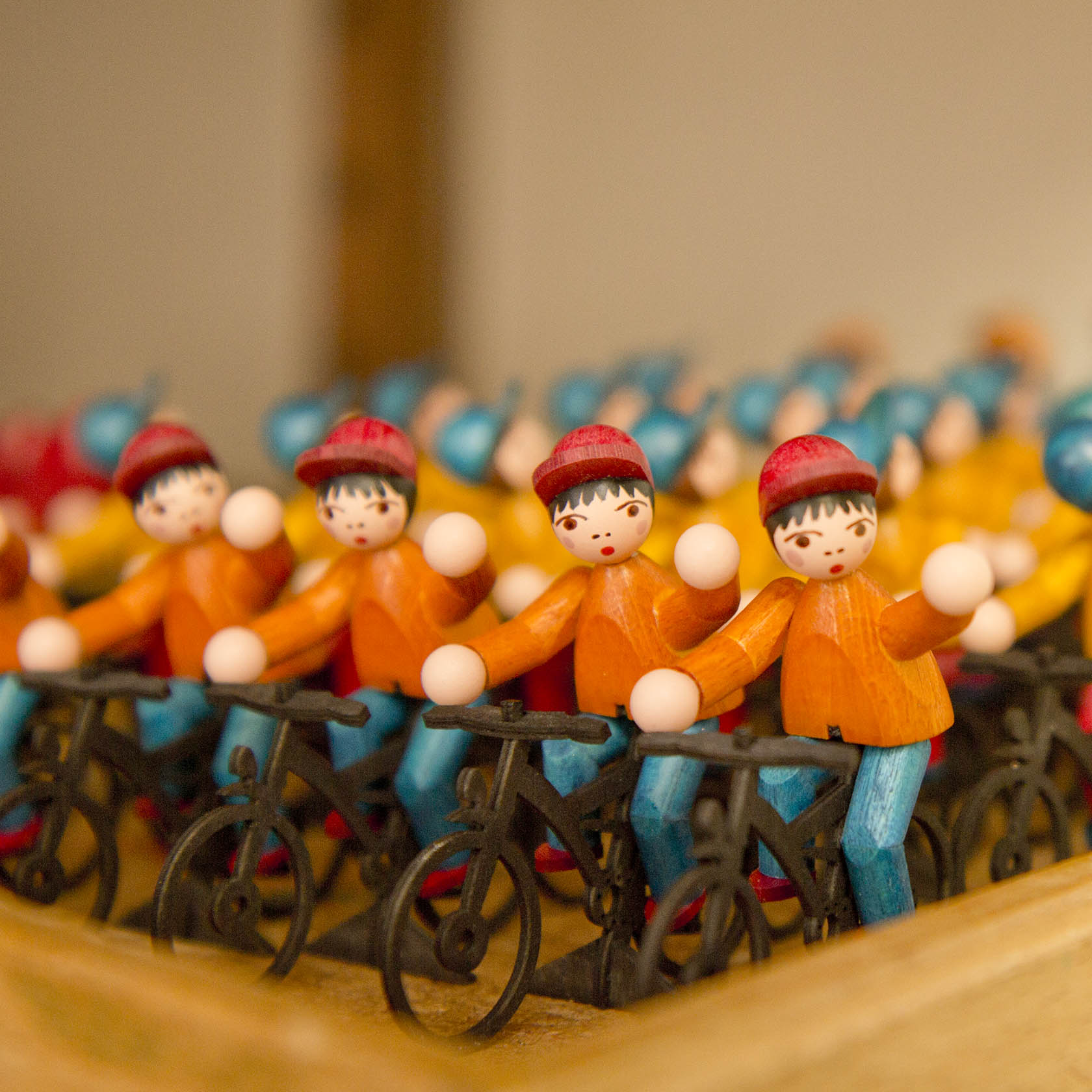 Holzfiguren in Form von Fahrradfahrern mit orangefarbenen Hemden und roten Mützen, hintereinander aufgestellt.