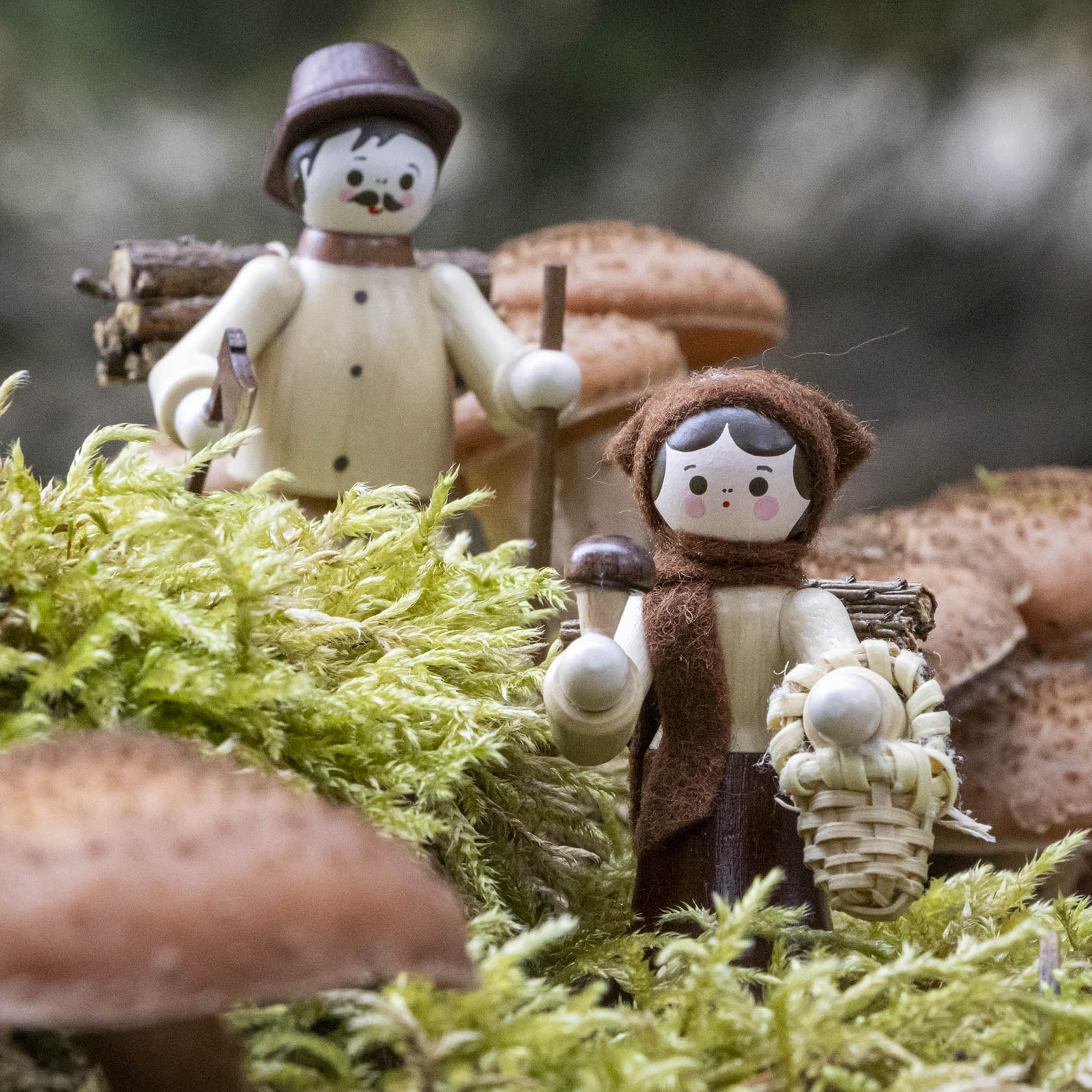 Holzfiguren eines Mannes und einer Frau in traditioneller Kleidung mit Wanderstöcken, umgeben von Moos und Pilzen.
