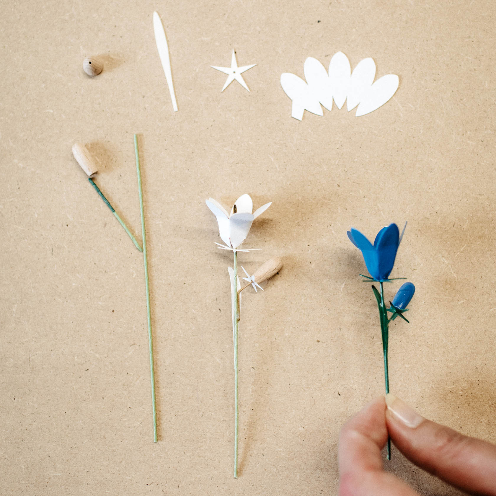 Eine Hand hält eine handgefertigte blaue Glockenblume mit Knospe aus Papier, daneben weitere Papierblumen und Einzelteile.