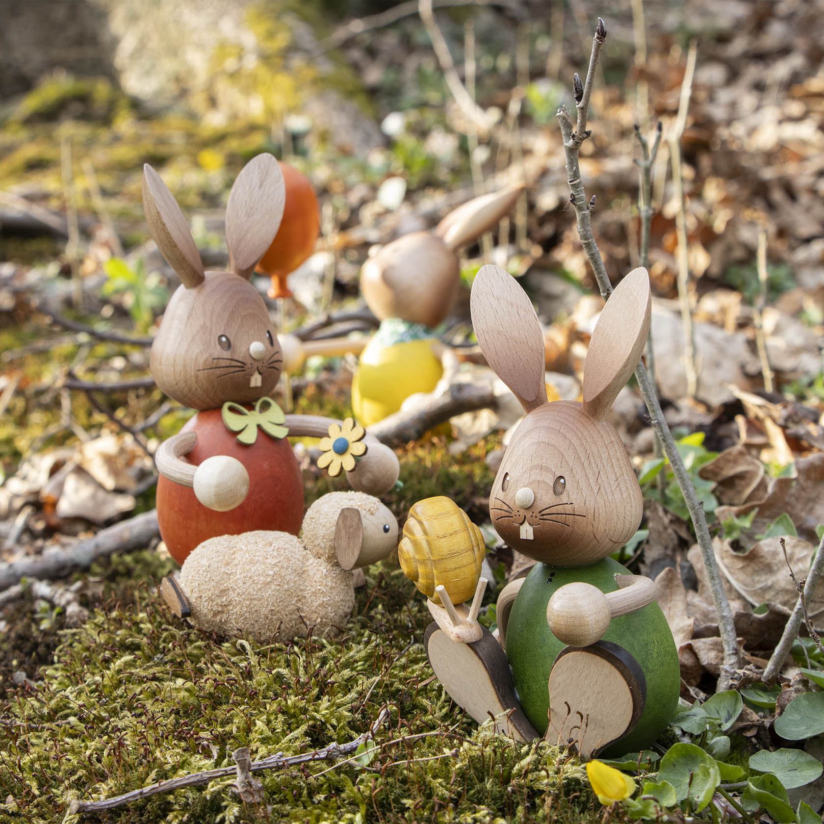 Holzhase mit orangefarbenem Pullover und grüner Hose, der auf Moos sitzt und ein gelbes Osterei hält, umgeben von weiteren Holzfiguren und Zweigen.