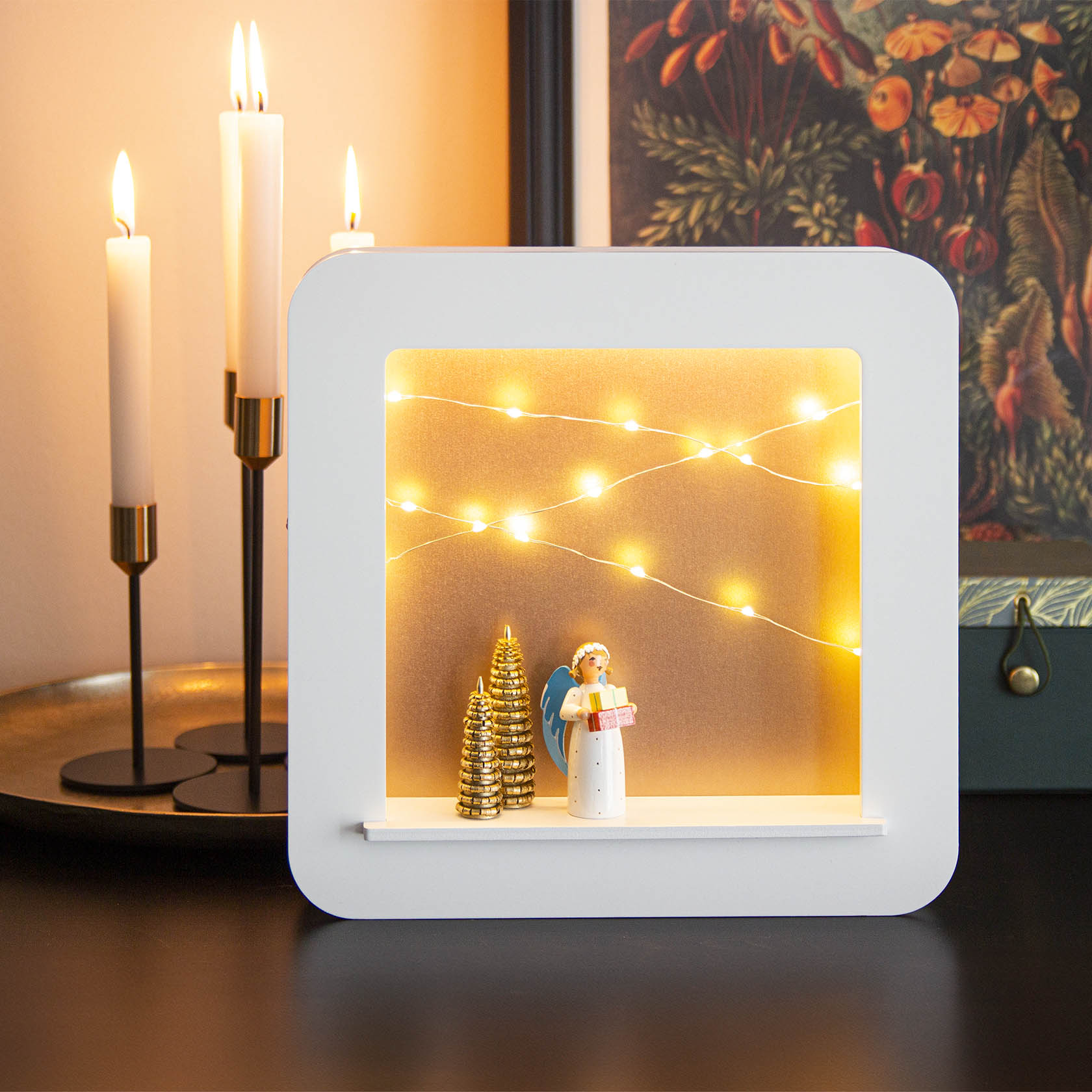 Weißer Objektrahmen mit LED-Beleuchtung, präsentiert eine Engelsfigur mit Geschenk und zwei goldenen Bäumchen. Im Hintergrund warme Lichter.