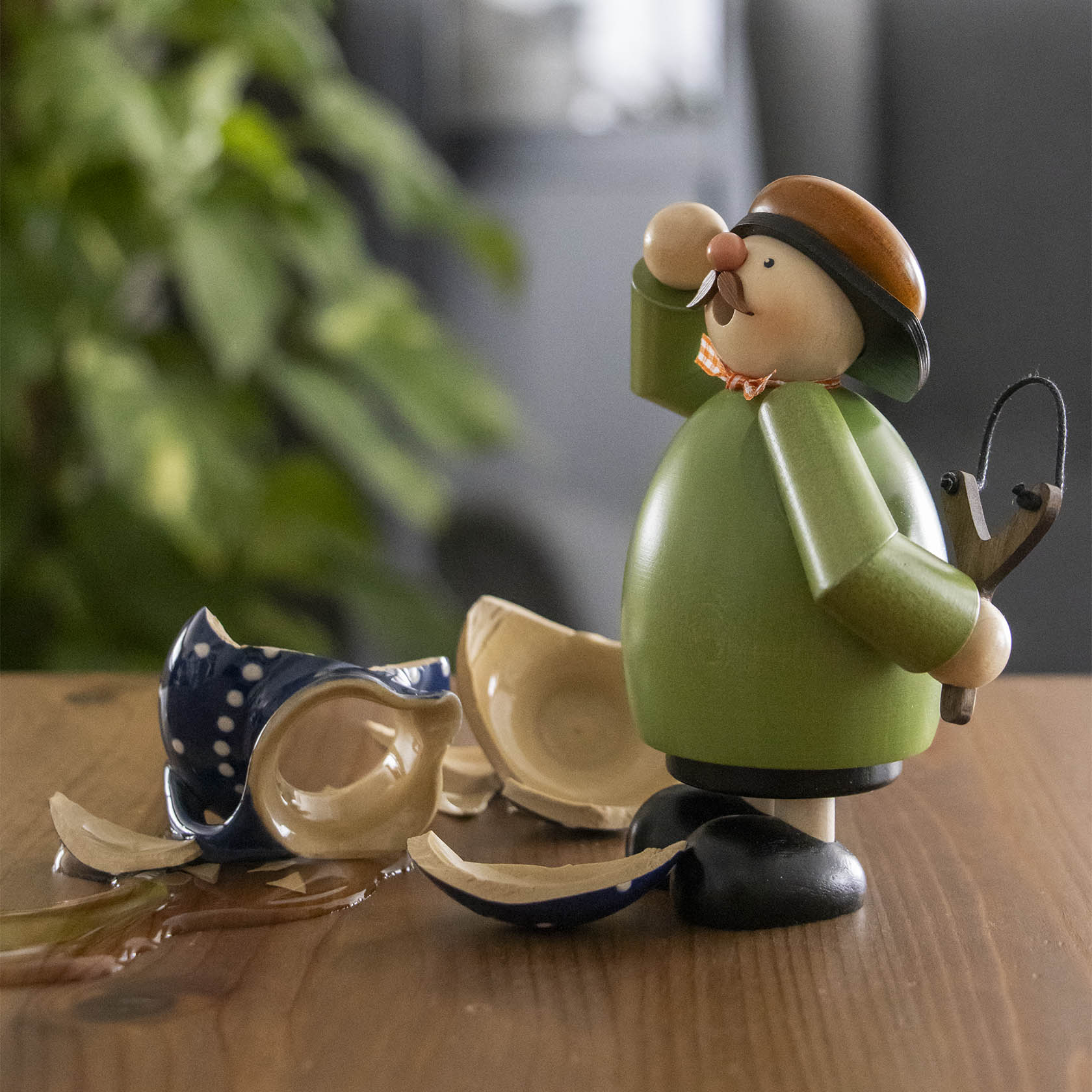 Ein Räuchermann in grüner Jacke und braunem Hut, hält eine Schleuder. Vor ihm liegt eine zerbrochene Vase.