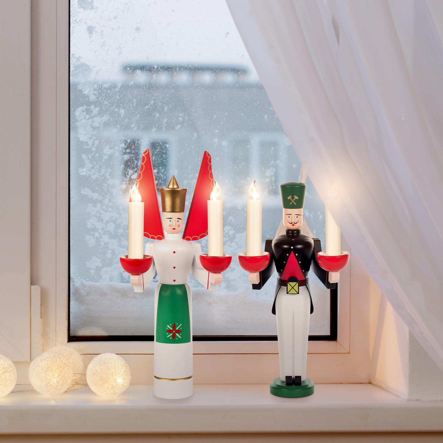 Dekorative Holzfiguren Engel und Bergmann auf einer Fensterbank. Der Engel trägt eine grüne Schürze und rote Flügel, beide Figuren halten Kerzen.