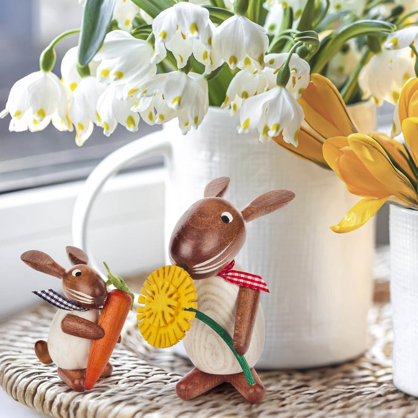 Zwei dekorative Holzhasenfiguren. Der größere Hase hält eine gelbe Blume, der kleinere eine orange Möhre. Beide tragen karierte Halstücher.