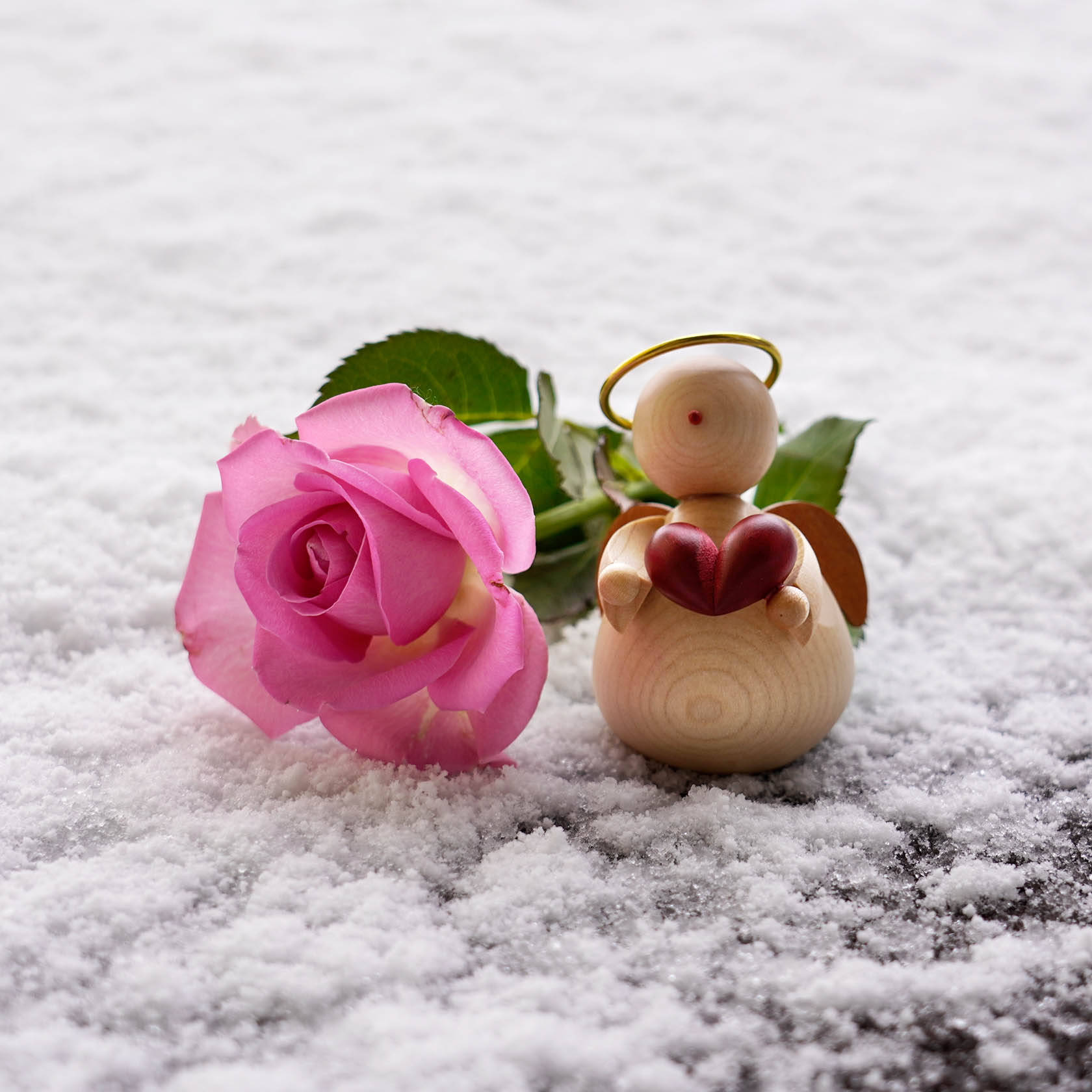 Ein Engel aus Holz mit einem roten Herz in der Hand steht auf schneebedecktem Boden neben einer rosa Rose.