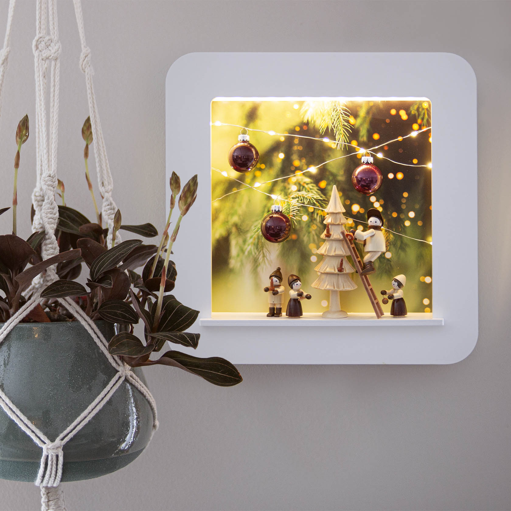 Weißer Objektrahmen mit LED-Beleuchtung zeigt eine weihnachtliche Szene mit Holzfiguren, einem Baum, roten Kugeln und Lichterketten.