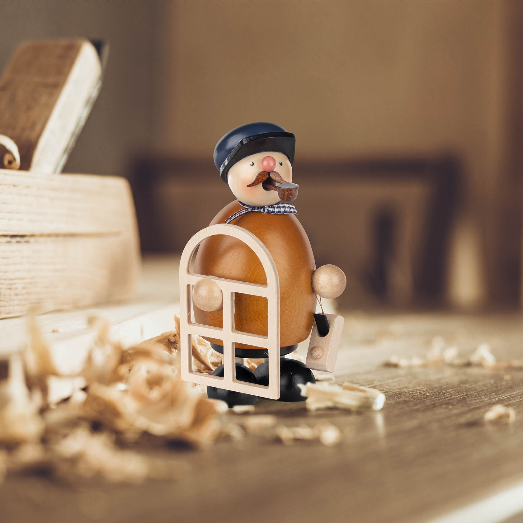 A traditional German incense smoker figure of a carpenter, with a blue cap, holding a window frame and surrounded by wood shavings.