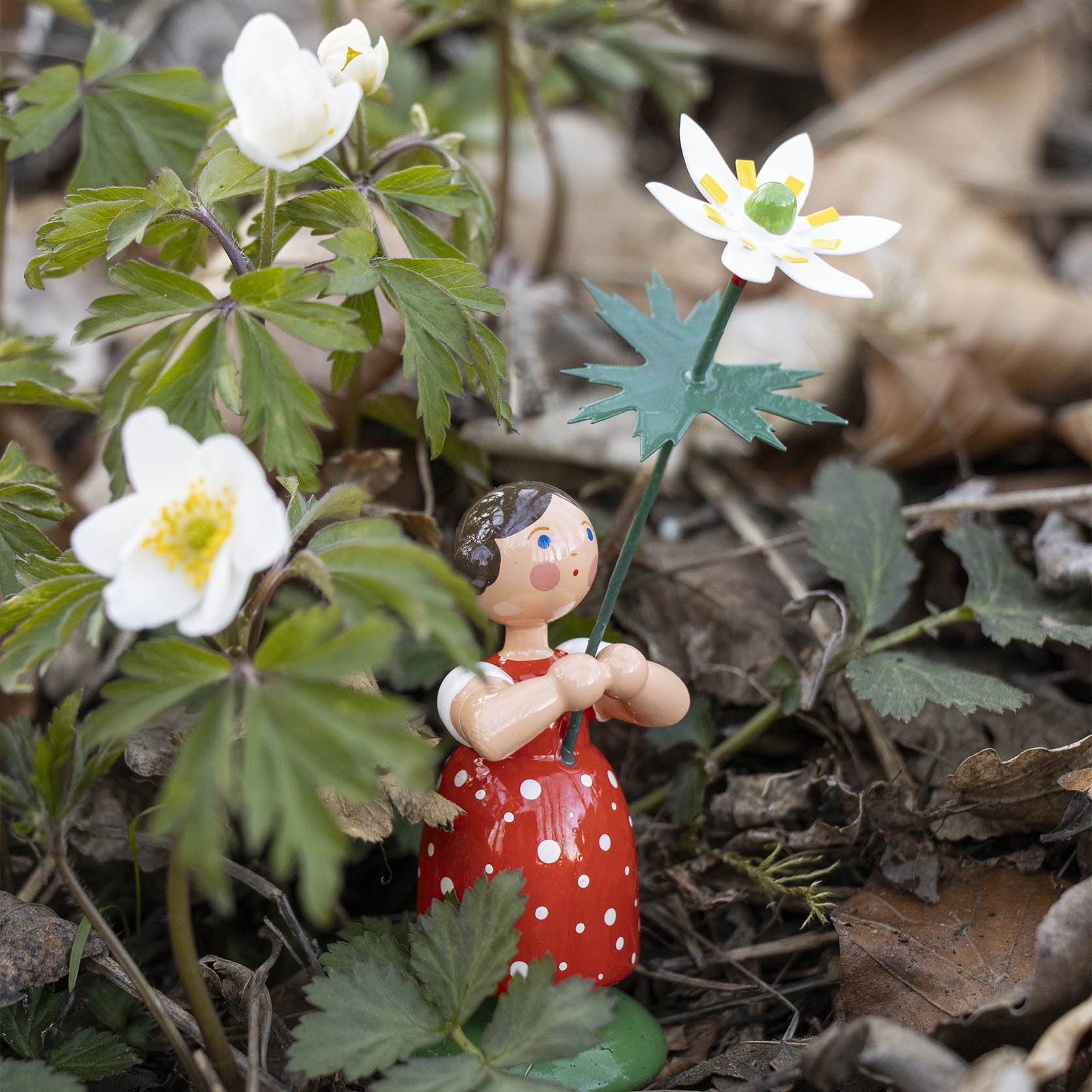 Holzfigur eines Mädchens in rotem Kleid mit weißen Punkten hält ein Buschwindröschen. Weitere Buschwindröschen umgeben die Figur.
