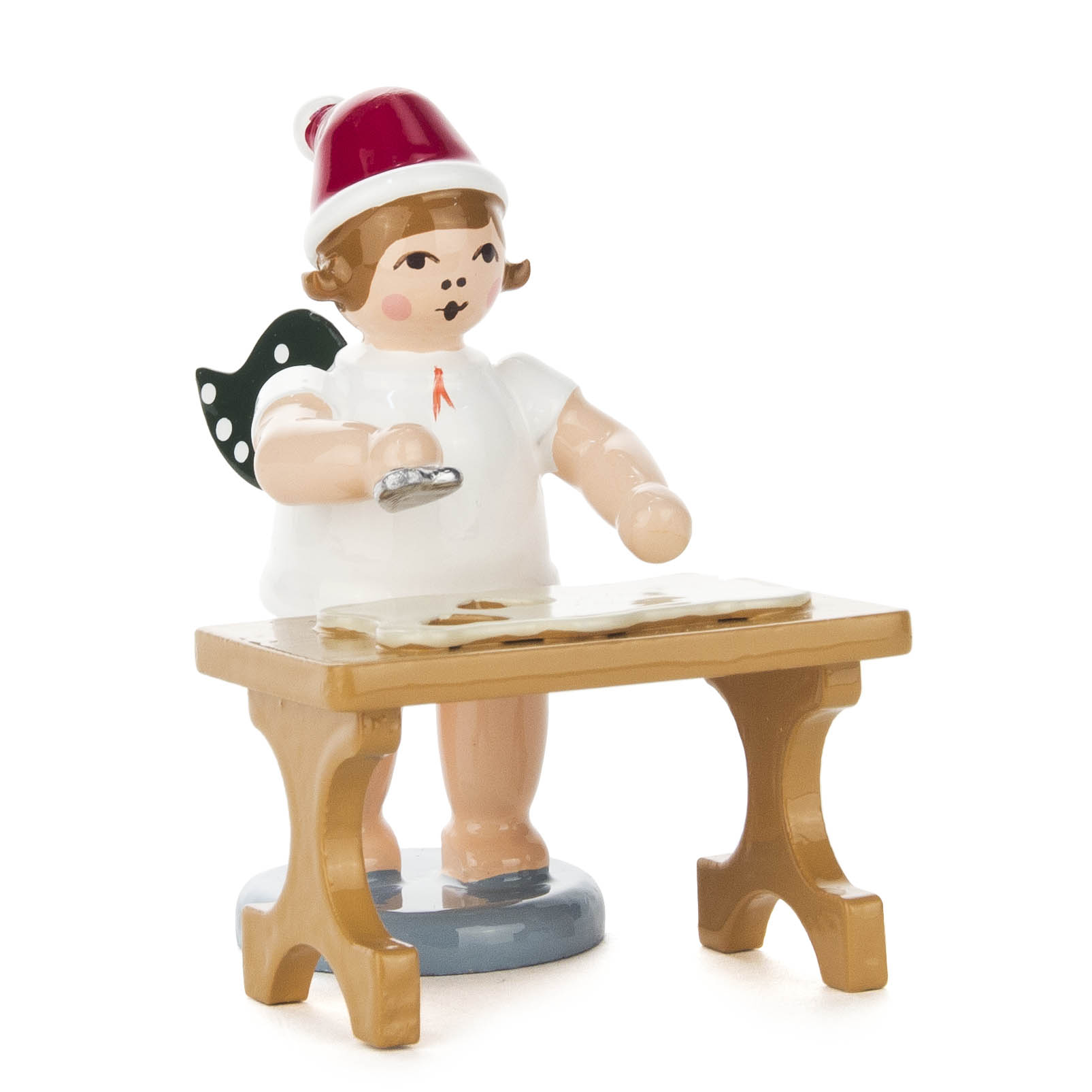 Ein Engel als Bäcker verkleidet mit grünen Flügeln und roter Weihnachtsmütze steht an einem Tisch und hält eine Ausstechform in der Hand. im Dregeno Online Shop günstig kaufen