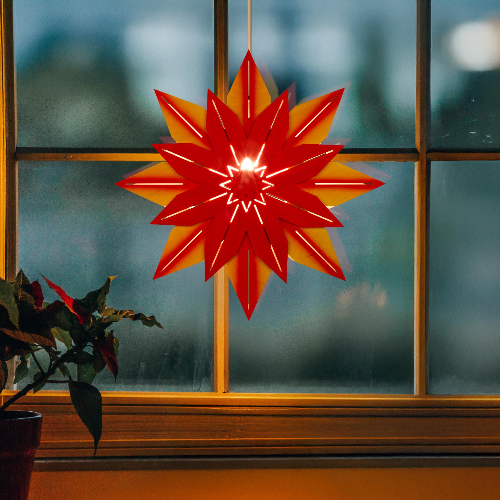 Leuchtend roter und gelber Weihnachtsstern hängt, mit elektrischer Beleuchtung in der Mitte, an einem Fenster.