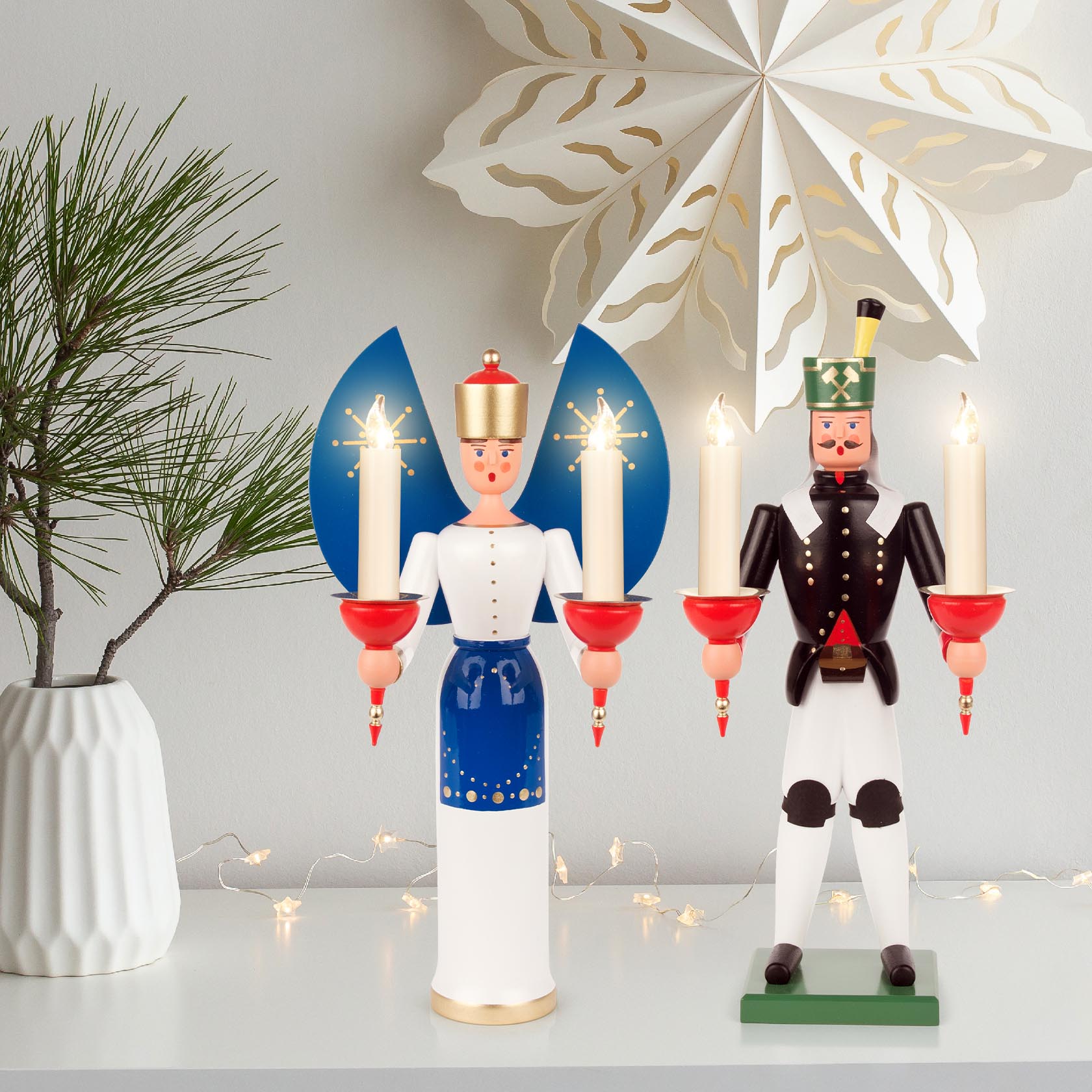 Holzengel mit blauen Flügeln, goldener Krone und zwei Kerzenhaltern, die brennende Kerzen tragen, neben einem Nussknacker mit Kerzen.