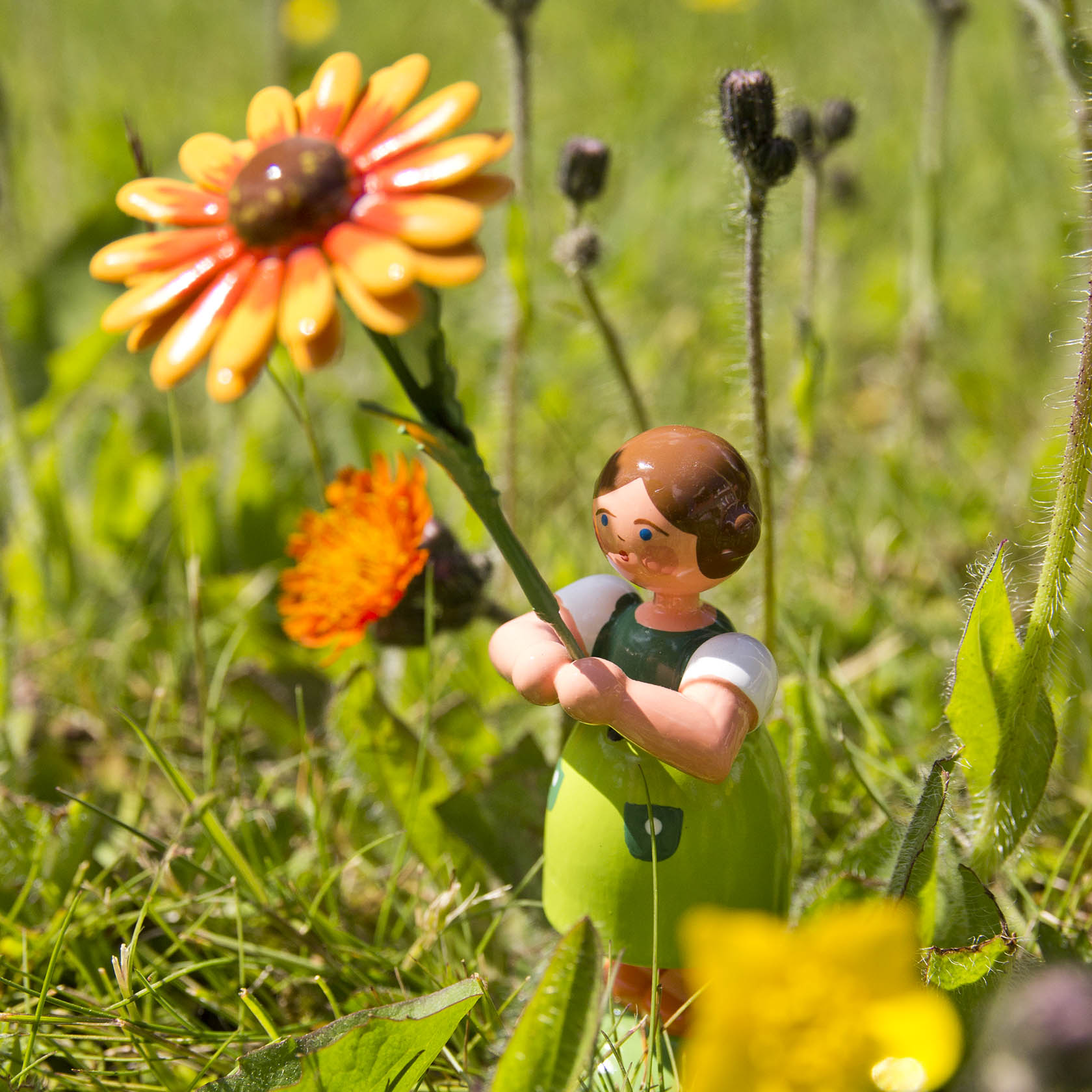 A small figurine of a girl holding a vibrant orange and yellow blanket flower in a grassy meadow.