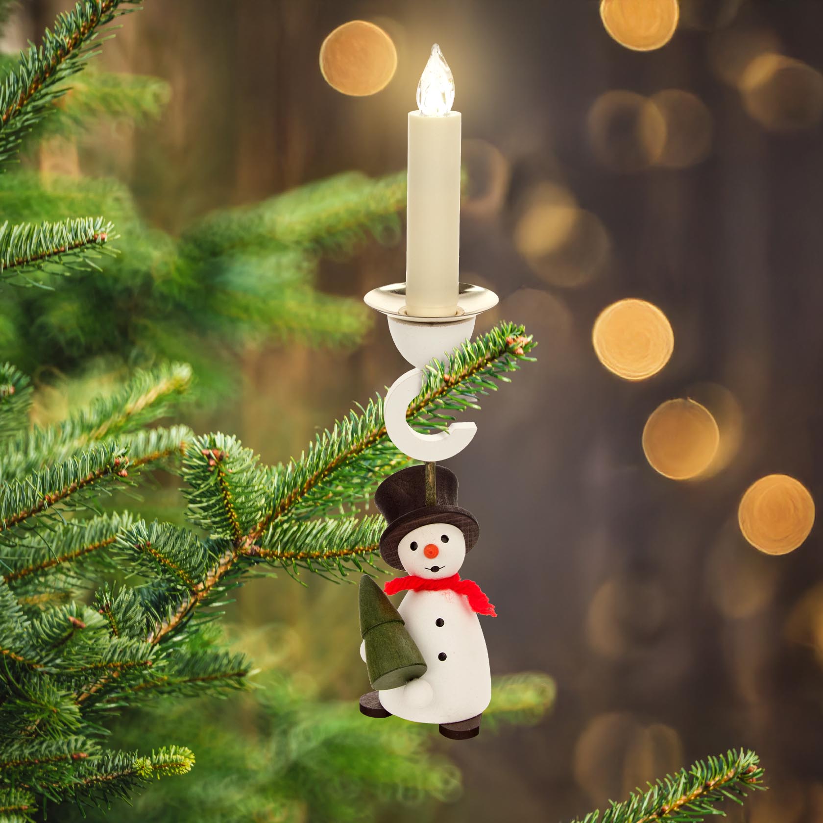 Ein Schneemann-Kerzenhalter mit schwarzem Zylinder und rotem Schal, der einen kleinen grünen Baum hält. Hängt an einem Tannenzweig, Kerze oben eingesteckt.