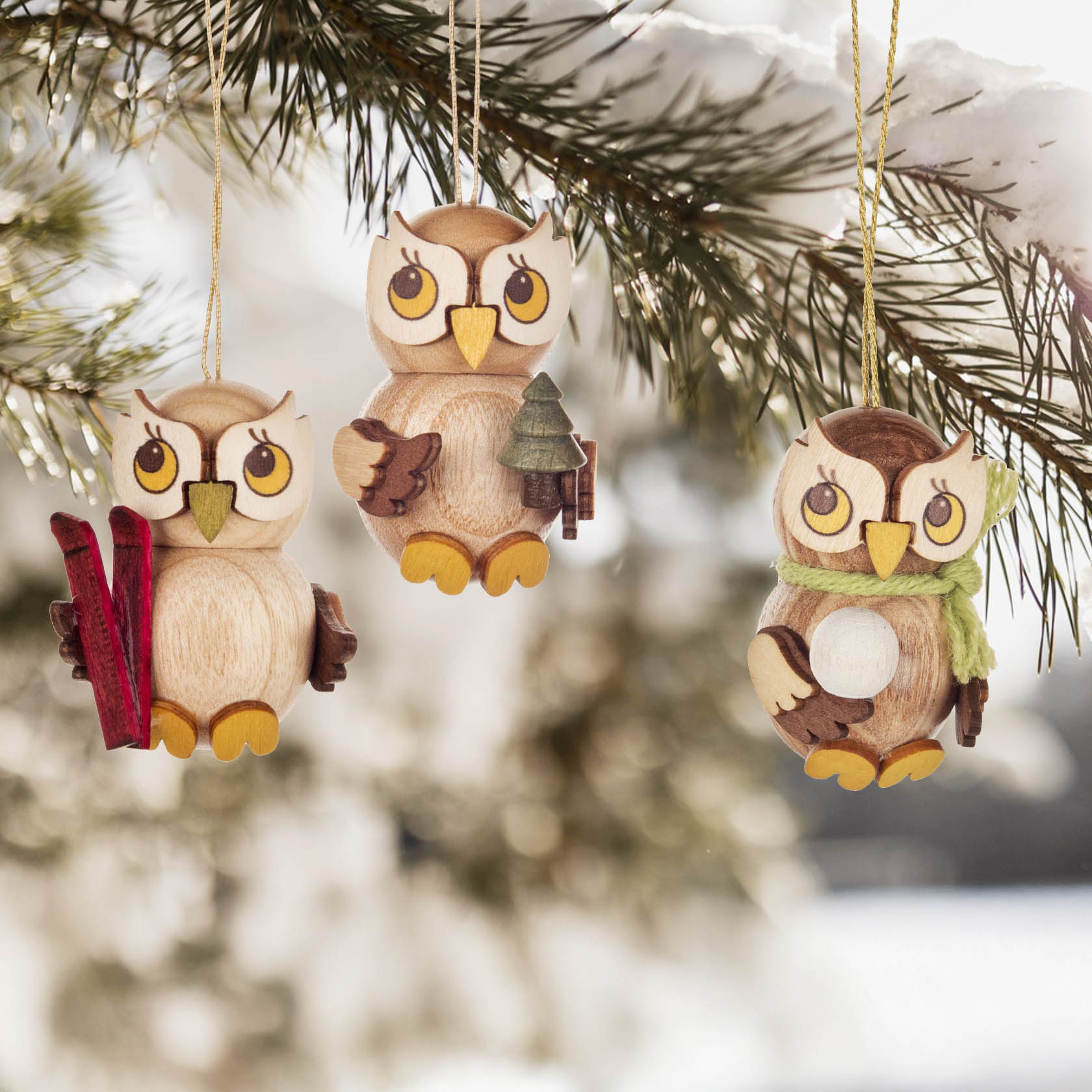 Drei hölzerne Eulen-Anhänger an einem Ast: links mit Skiern, in der Mitte mit einem Baum, rechts mit einem Schneeball.