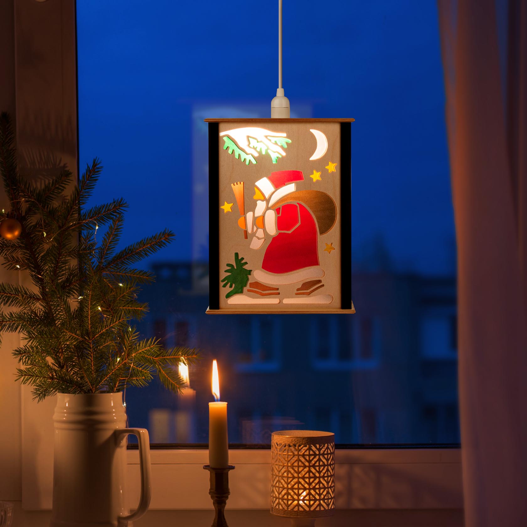 Eine beleuchtete Fensterlaterne mit einem Weihnachtsmannmotiv, die an einem Fenster hängt. Ein Weihnachtsbaum steht daneben und eine Kerze brennt vorne.