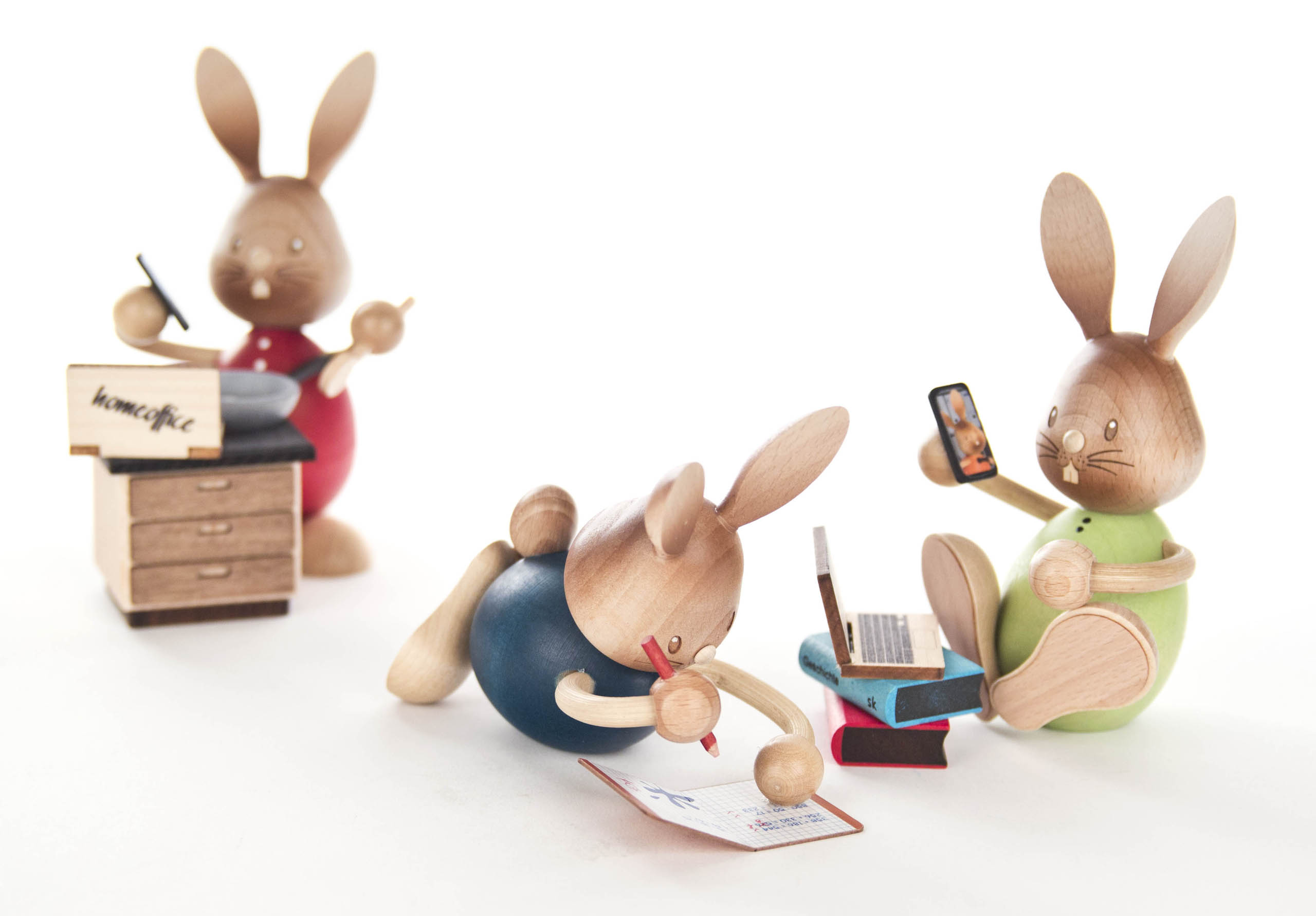 Three wooden bunny figures engaged in different activities: one writing in a notebook, another on a laptop with a smartphone, and a third at a desk.