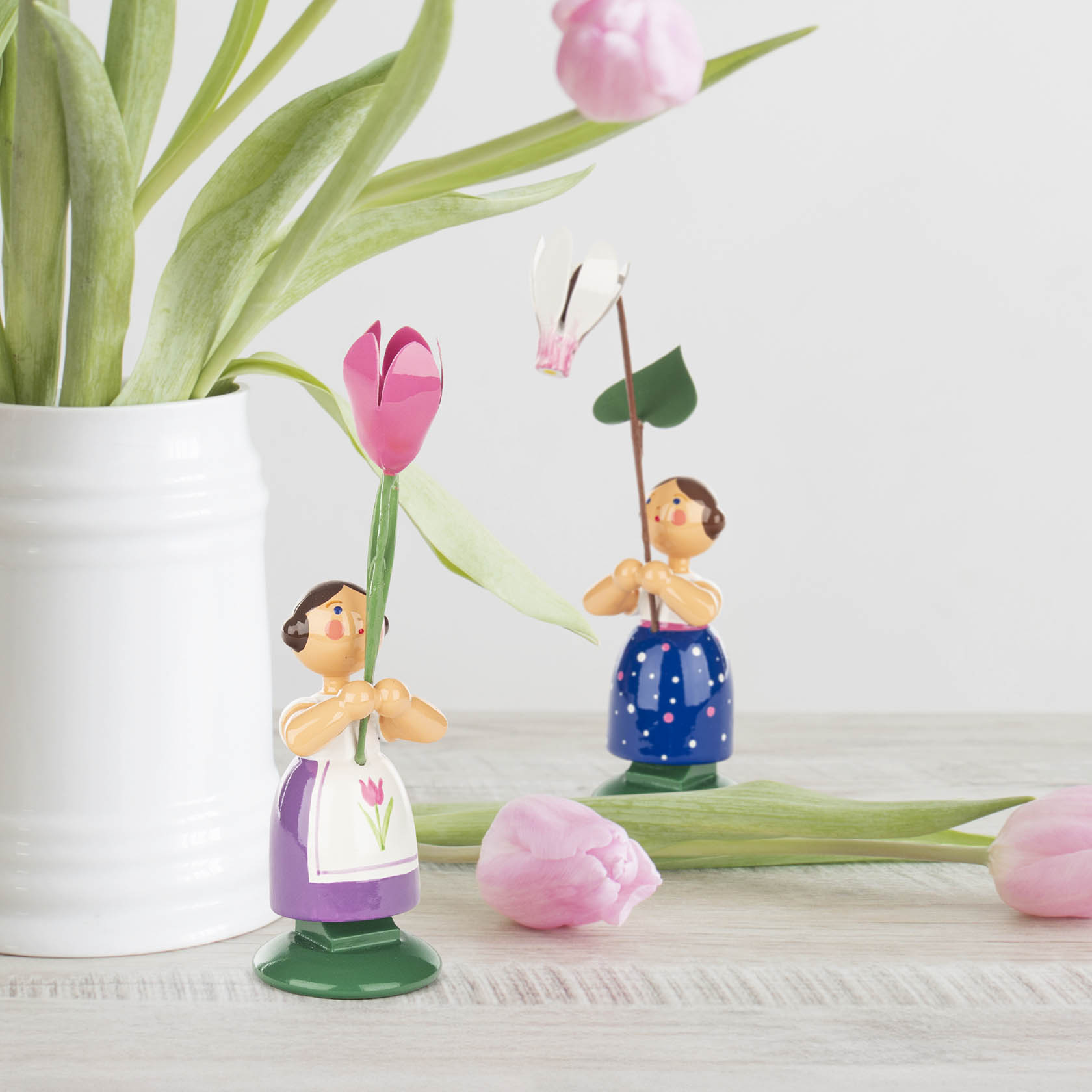 Zwei Blumenmädchen-Figuren aus Holz, eine mit rosafarbenem Alpenveilchen, die andere in blauer Kleidung mit rosa Punkten, auf einem hellen Tisch.