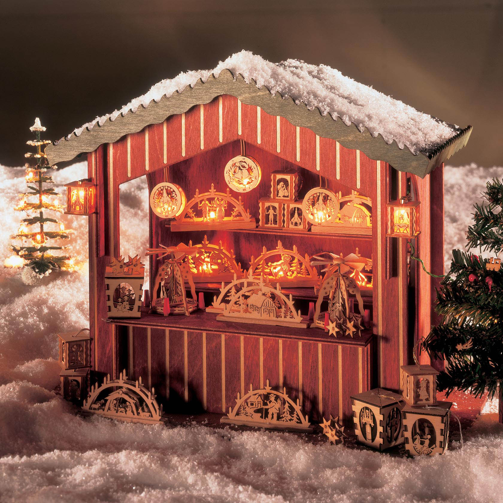 Holz-Weihnachtsmarktstand mit leuchtenden Schwibbögen und Ornamenten, umgeben von künstlichem Schnee und einem kleinen beleuchteten Baum.