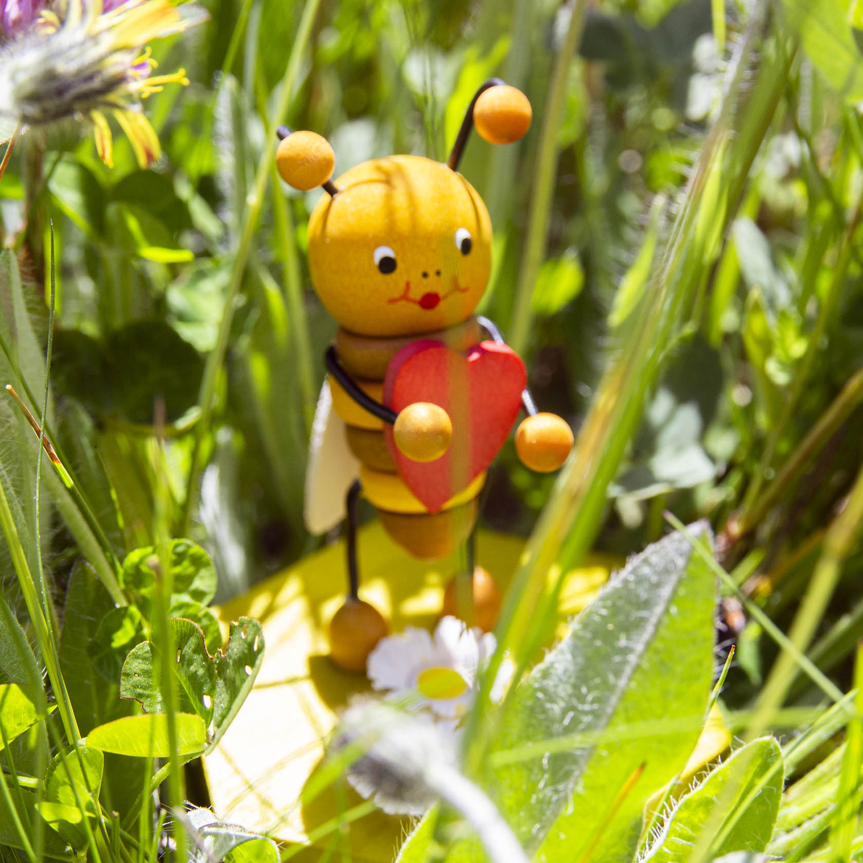 Eine hölzerne Biene hält ein rotes Herz in den Armen, umgeben von grünen Grashalmen und einer weißen Blume.