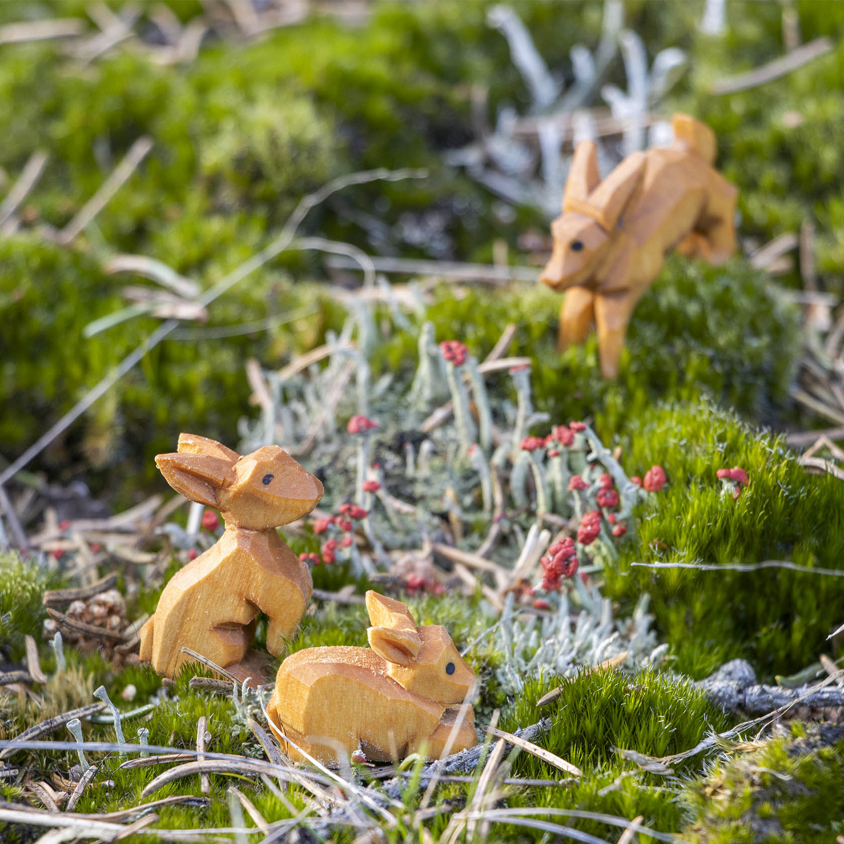 Drei handgeschnitzte Holzhasen auf moosbewachsenem Boden mit kleinen roten Blumen. Zwei sitzen nebeneinander, der dritte steht im Hintergrund.