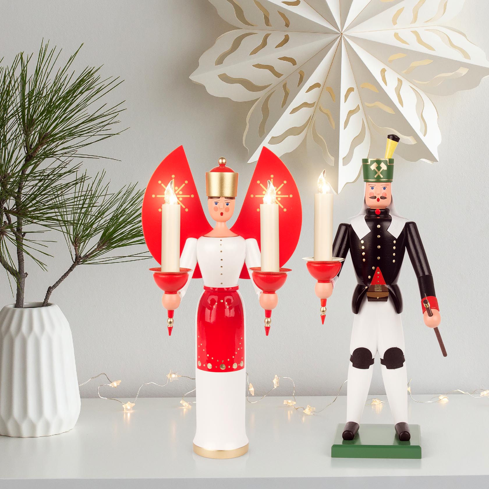 Ein traditioneller Erzgebirge-Bergmann aus Holz in Schwarz-Weiß, hält einen Kerzenhalter mit glimmender Kerze. Daneben ein strahlender roter Weihnachtsengel.