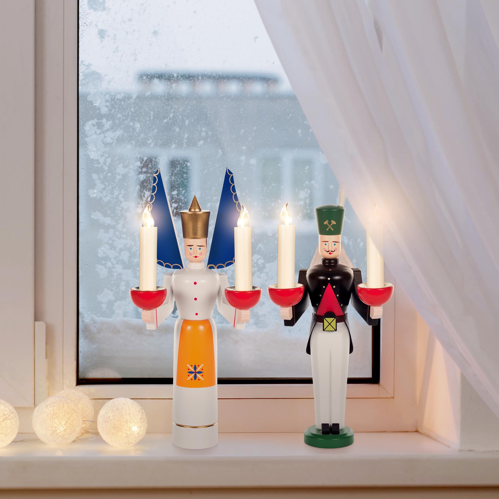 A wooden angel figurine with a golden crown, orange apron, and blue wings holds two lit candles. Next to it, a wooden miner figure with a green hat holds two lit candles.