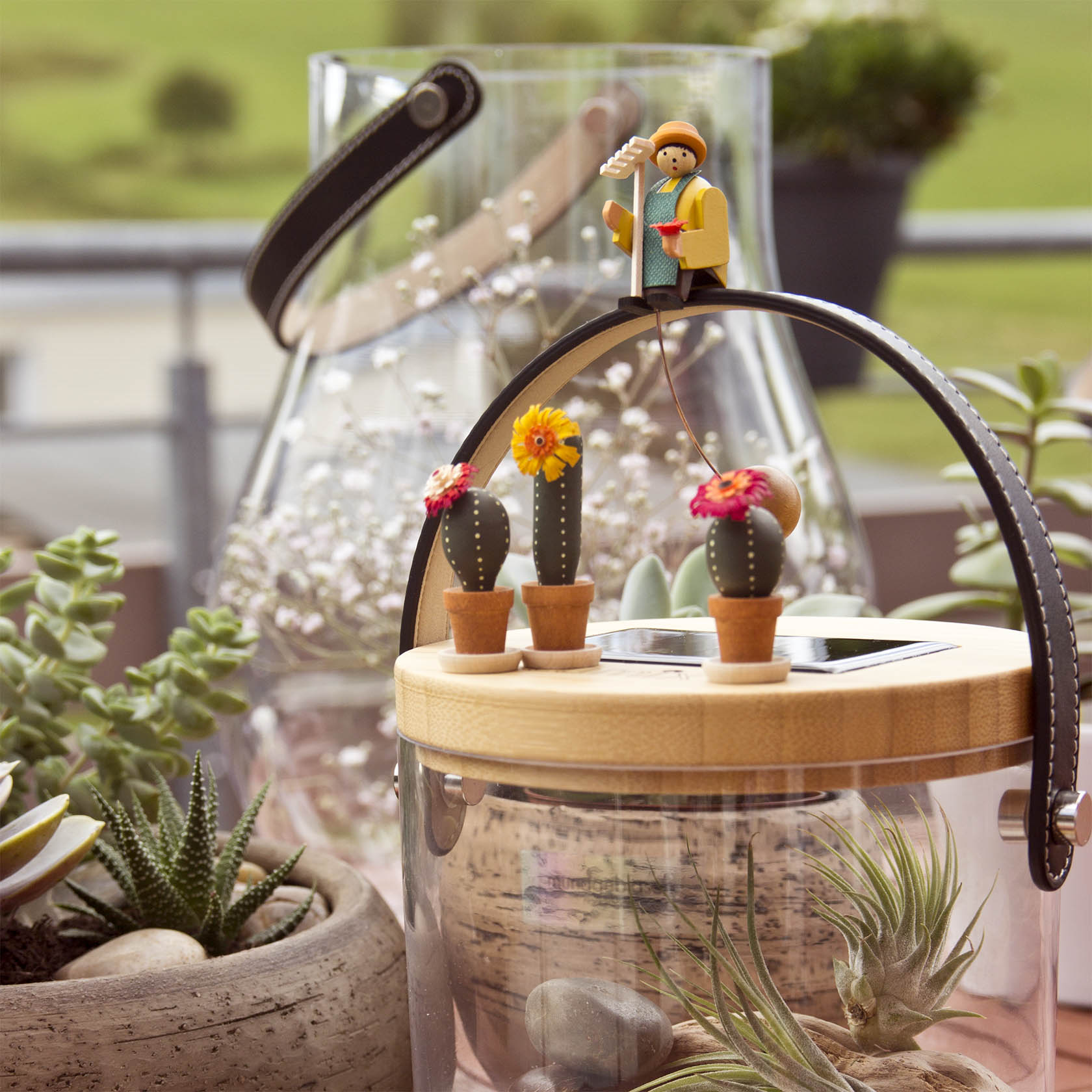 Eine Miniaturfigur eines Gärtners mit Spaten und Gießkanne steht auf einem Bogen über einem Terrarium. Daneben stehen kleine Modelle von Kakteen.