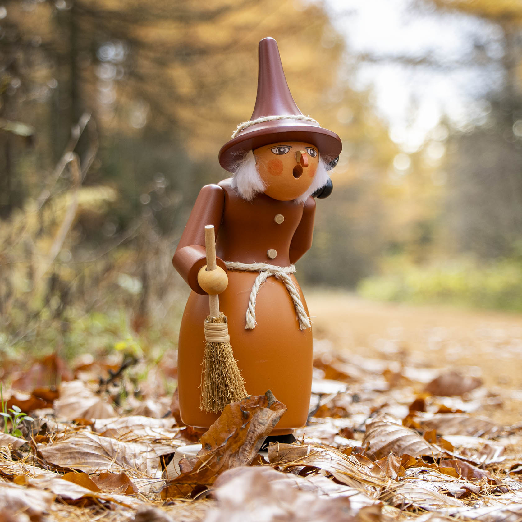 Wooden smoking figure of a witch with a tall hat, white hair, and a broom, standing on a path surrounded by autumn leaves.