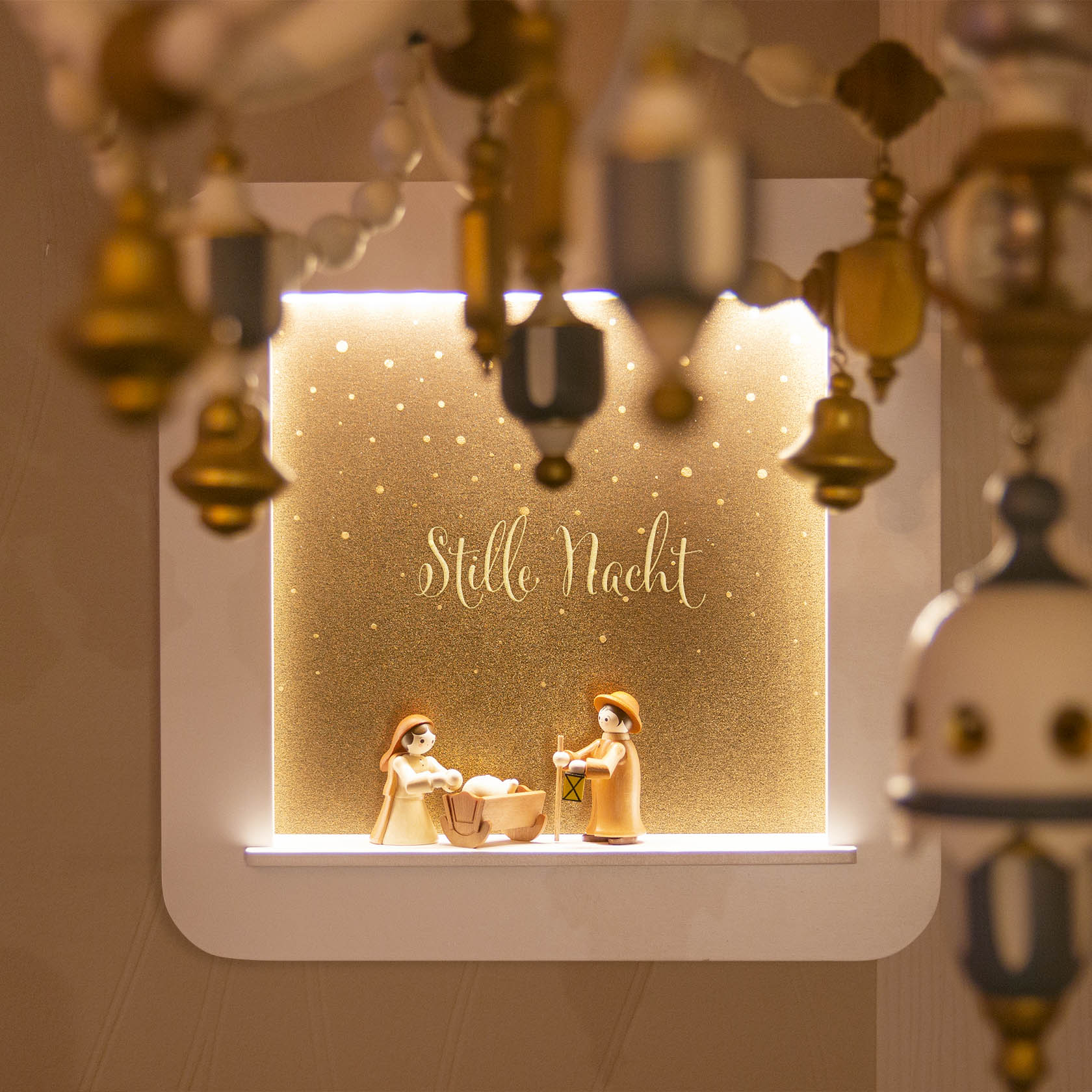 Ein beleuchteter Objektrahmen zeigt eine Krippenszene mit Maria, Josef und Jesuskind. Der Hintergrund mit dem Schriftzug 'Stille Nacht' ist in goldenem Schimmer gehalten.