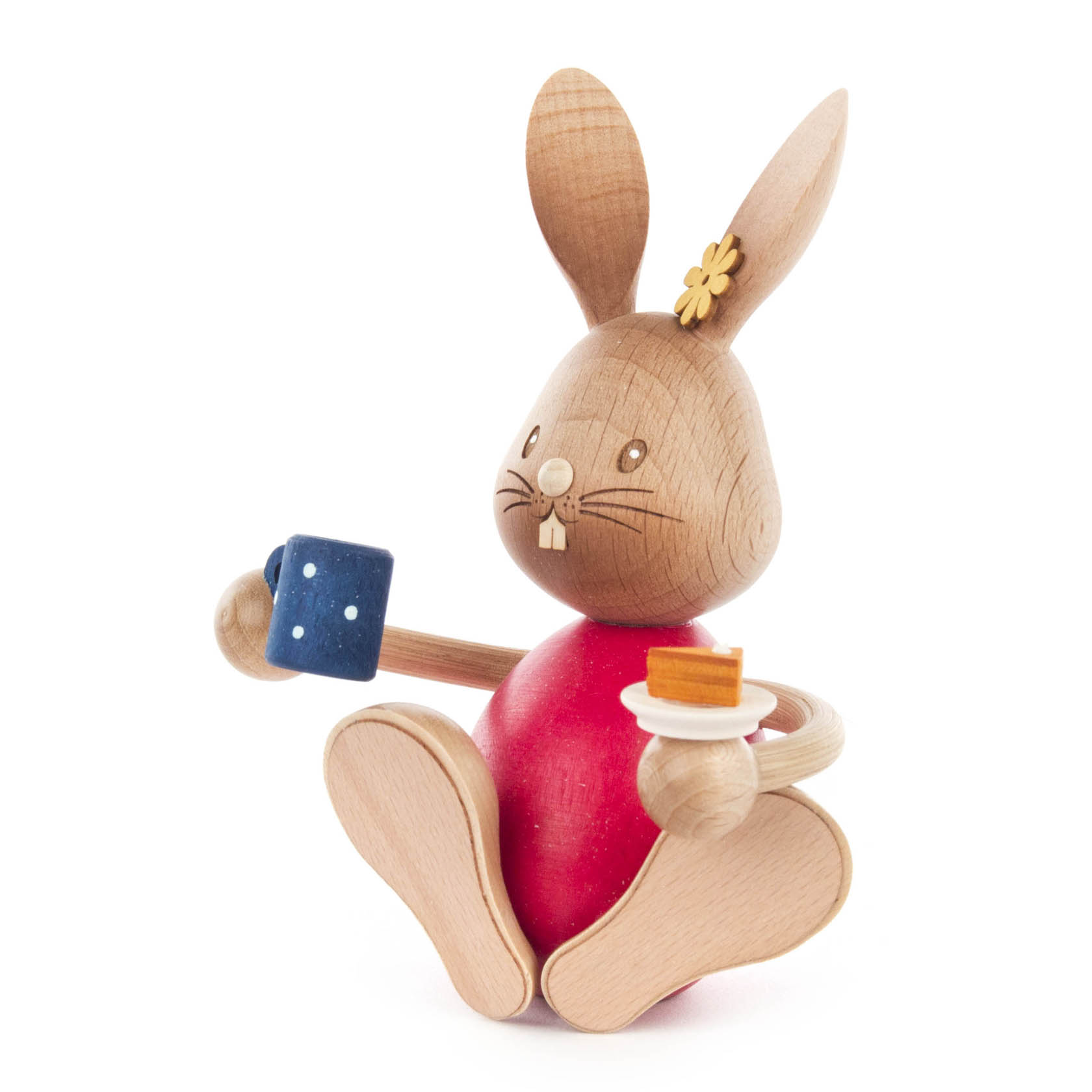 Holzhase Stupsi sitzt mit einer blauen Kaffeetasse in der linken Hand und einem Kuchenteller mit Kuchenstück in der rechten Hand.