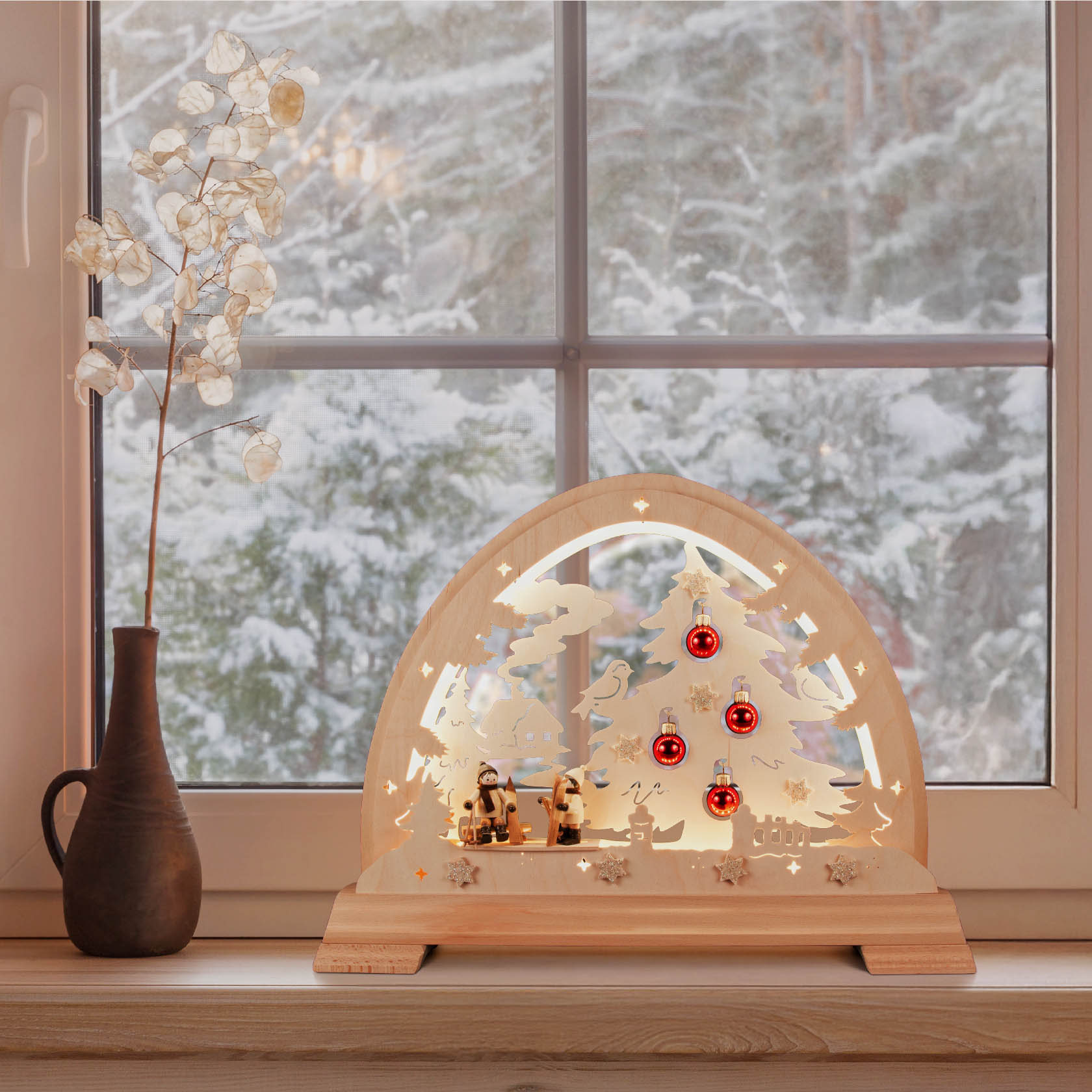 Ein beleuchteter Holz-Rundbogen mit Skifahrern, einem Weihnachtsbaum mit roten Kugeln und einer LED-Beleuchtung steht auf einem Fensterbrett.
