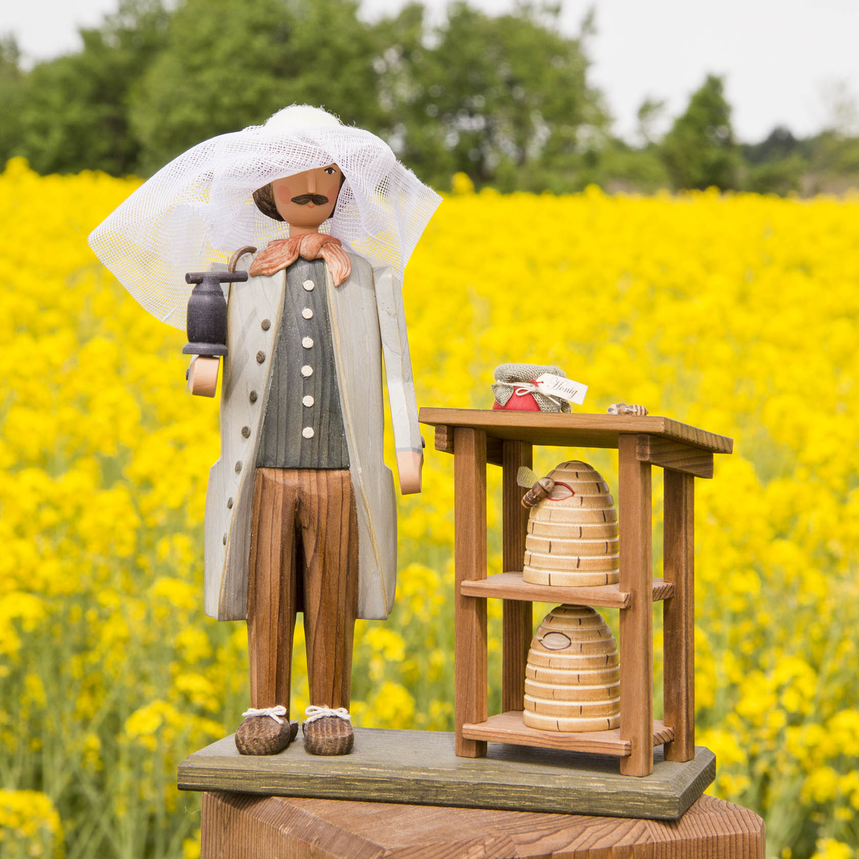 Holzfigur eines Imkers mit Schleierhut, in der linken Hand eine Kanne haltend, neben zwei Bienenkörben auf einem kleinen Holztisch.