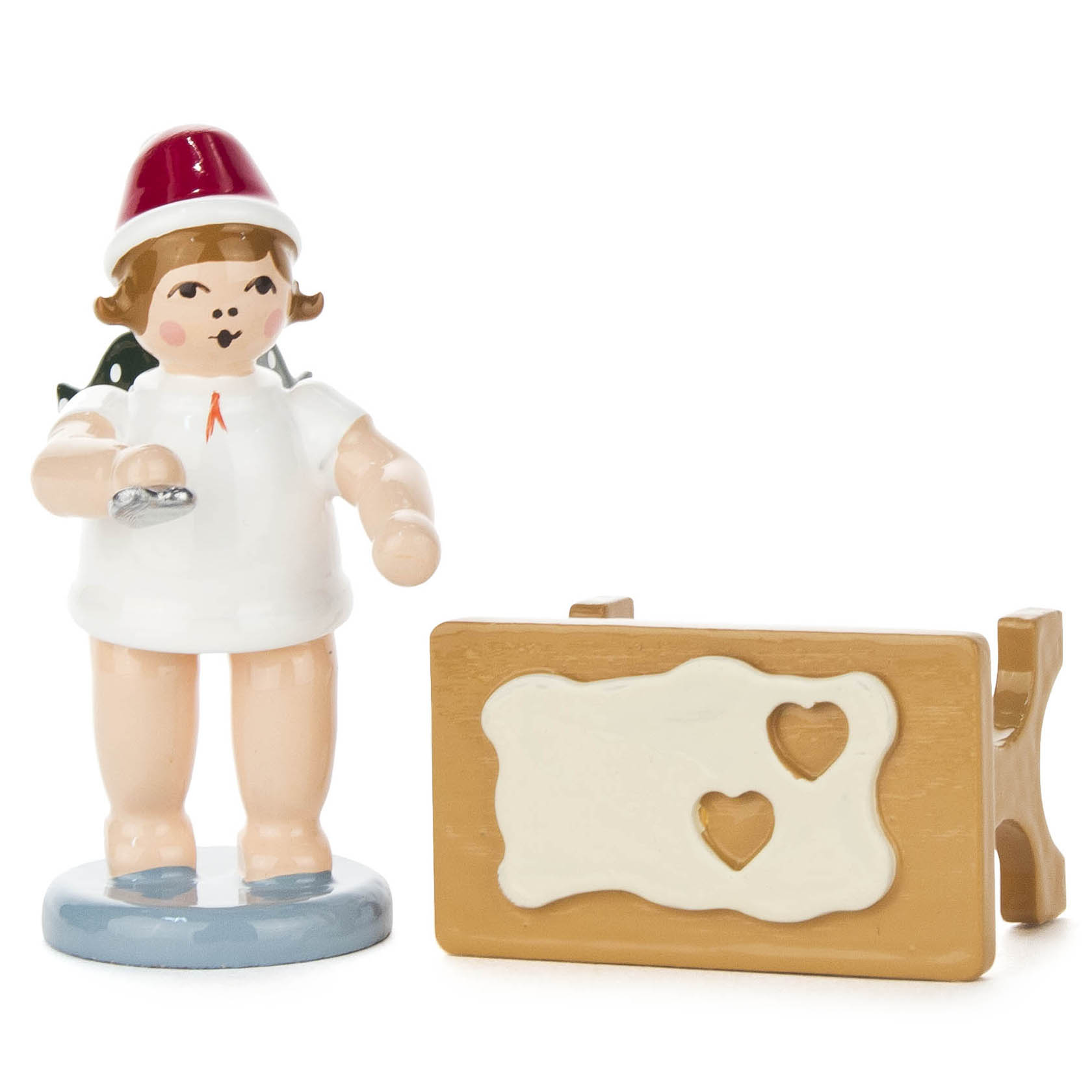 Figur eines Bäckerengels mit Mütze und einer Ausstechform in der Hand. Daneben ein Tisch mit einem Teigstück und zwei ausgestochenen Herzen.