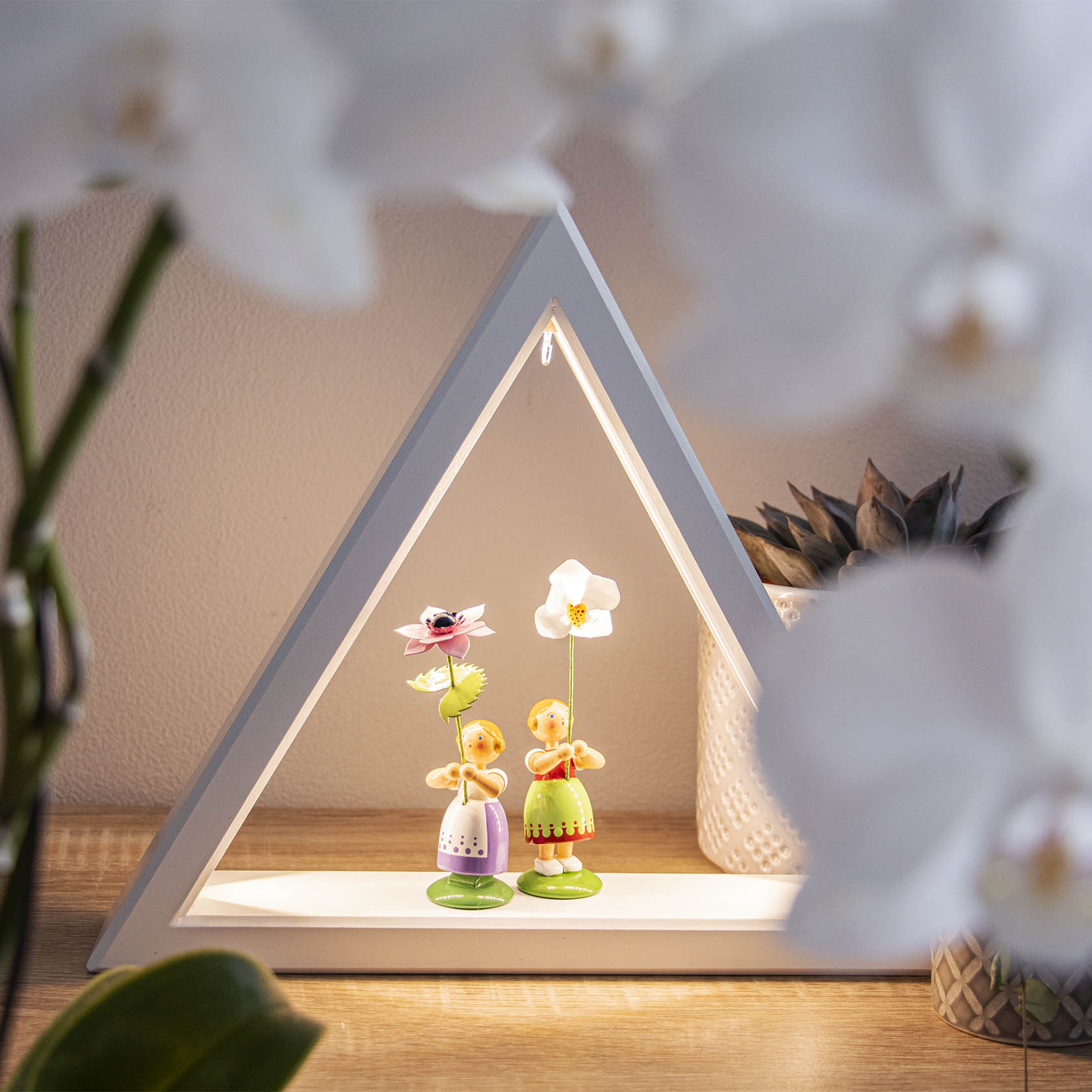 Zwei Holzfiguren mit Blumen, die Mädchen darstellen. Das linke Mädchen hält eine rosa Anemone, das rechte eine weiße Blume. Sie stehen in einem dreieckigen Regal.
