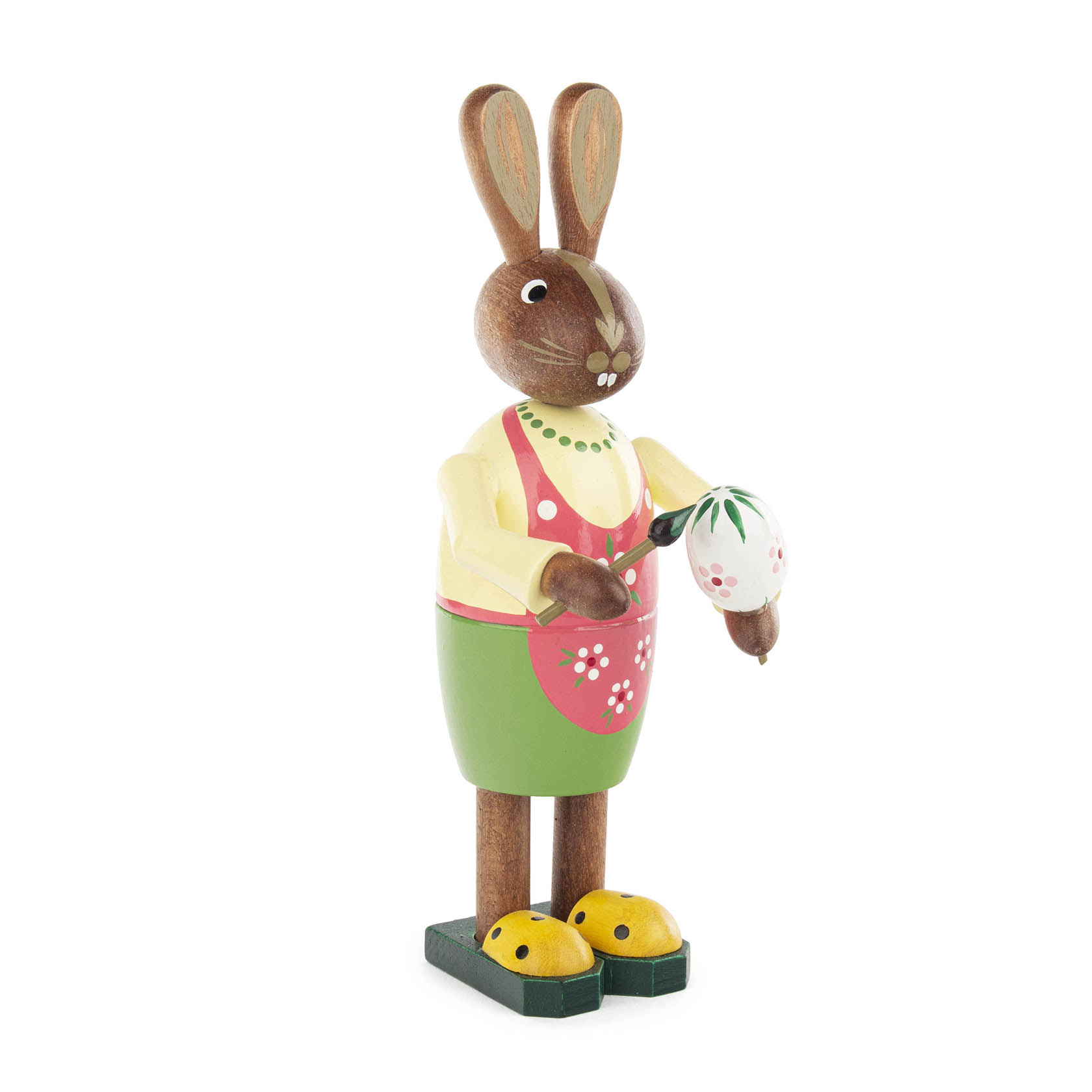 Holzfigur einer Osterhasenfrau, die ein Osterei bunt bemalt. Sie trägt eine grüne Schürze mit pinkem Schurz, gelben Pullover und gelbe Schuhe. im Dregeno Online Shop günstig kaufen