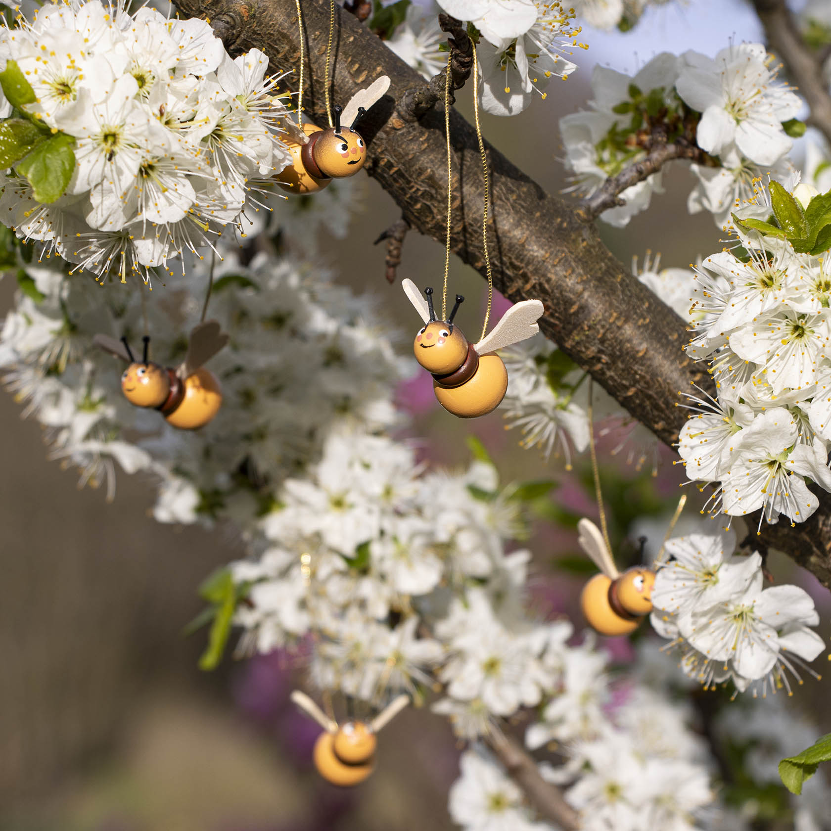 Fünf kleine, hängende Dekorationsobjekte in Form von Hummeln an einem blühenden Ast eines Obstbaums mit weißen Blüten.