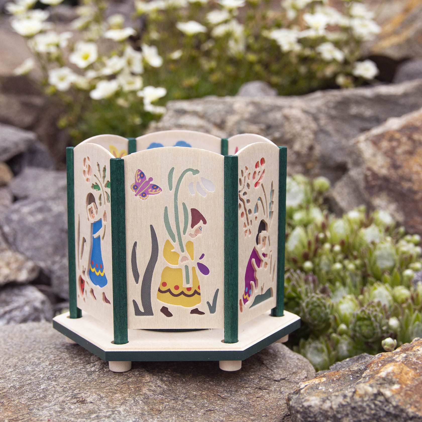Ein hexagonaler Teelichthalter aus Holz mit ausgeschnittenen Blumenkindmotiven. Kind mit Schneeglöckchen, Kind mit Mohnblumen und Kind mit süßer Blume sind zu sehen.