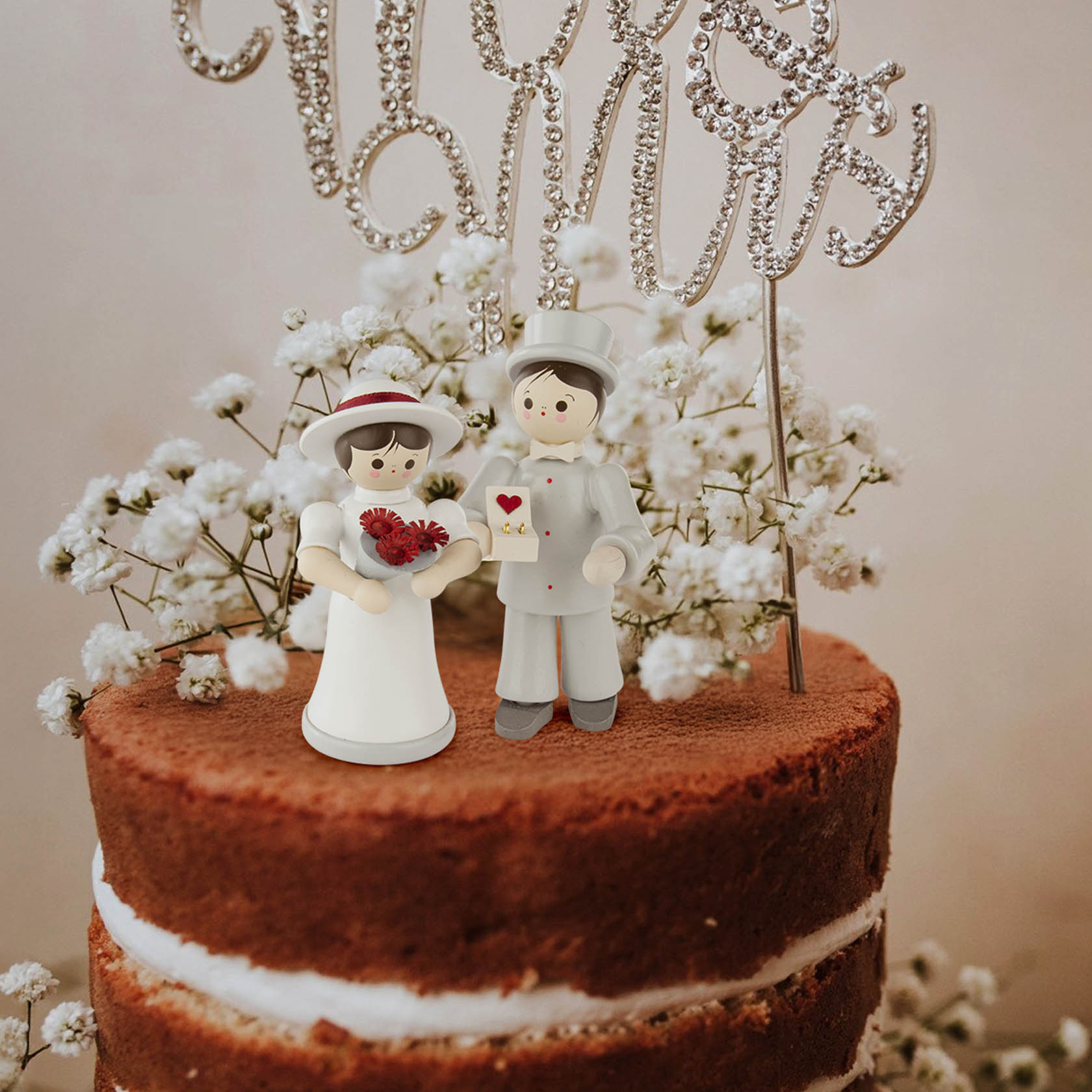 Figuren eines Brautpaars aus Plastik auf einer Hochzeitstorte. Die Braut trägt einen weißen Hut und hält einen Blumenstrauß, der Bräutigam einen Brief.