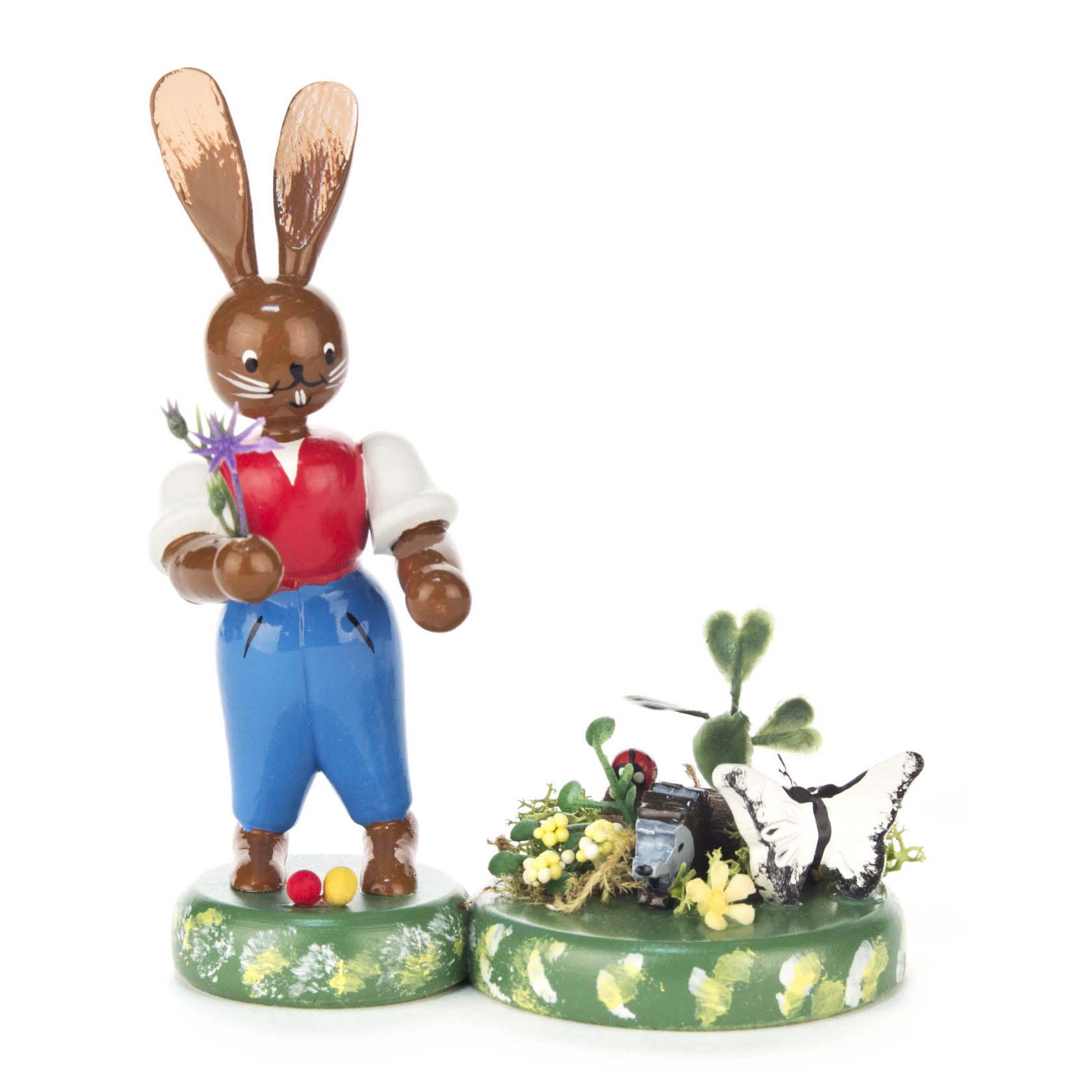 Hasenfigur in blauer Hose und rotem Hemd, die eine Blume hält, steht neben einer Frühlingsszene mit gelben Blumen, einem Schmetterling und Osterdekorationen. im Dregeno Online Shop günstig kaufen