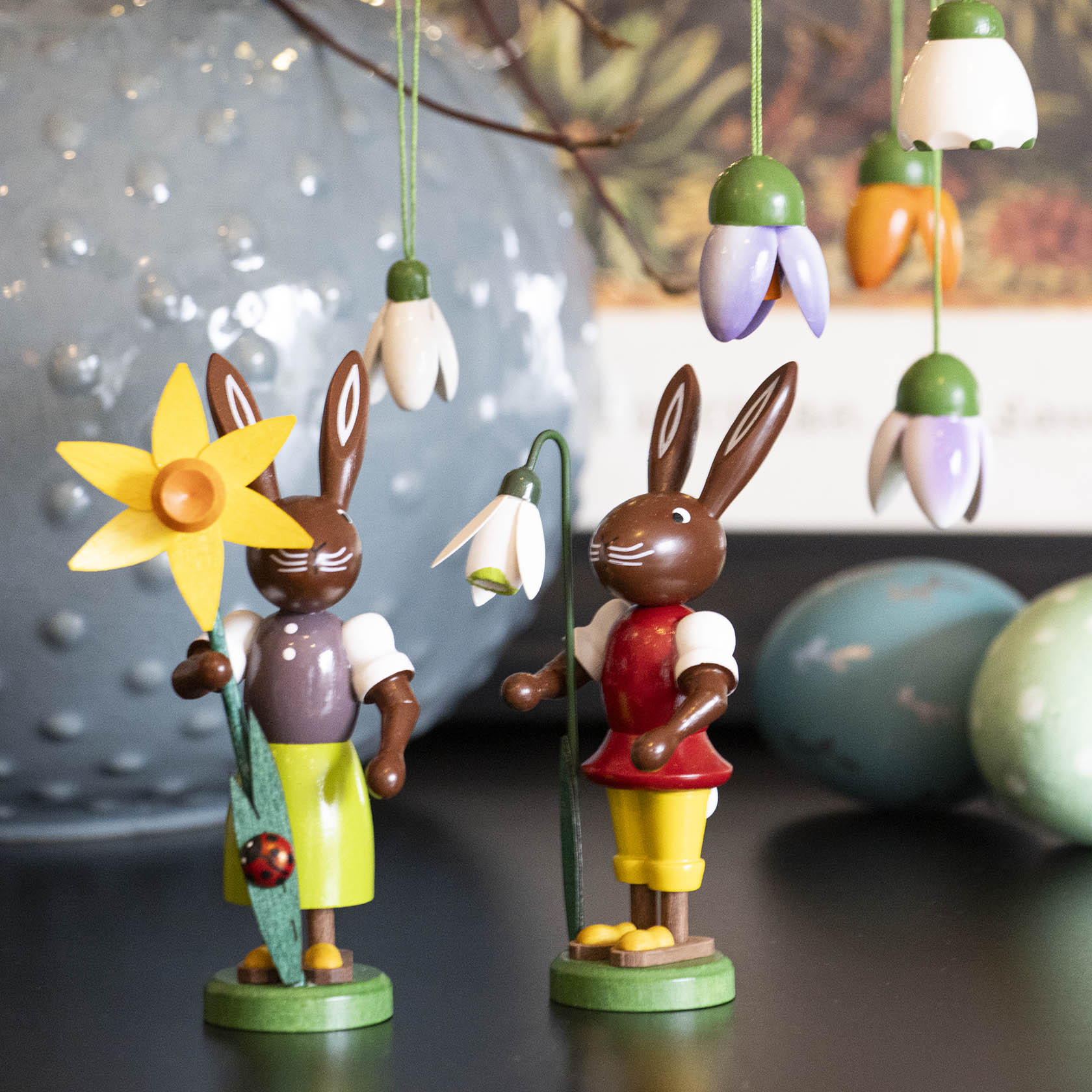 Zwei dunkle Holzhasenfiguren mit Frühlingsblumen und bunter Kleidung. Hängebunte Frühlingsblumen und ein hellblaues Ei sind im Hintergrund.