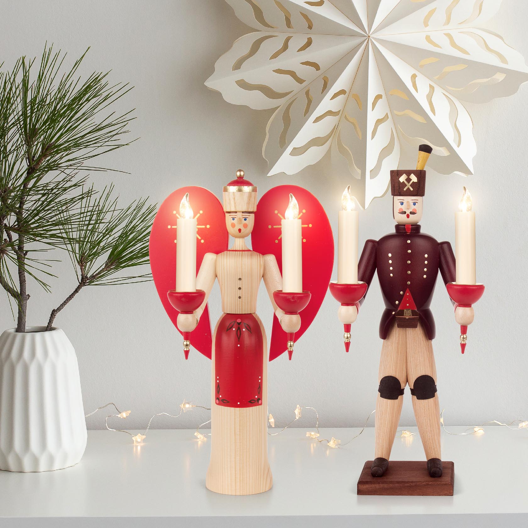 Two wooden candle holders shaped like miners. The left is painted red and beige with a heart-shaped background and three candles. The right is natural and brown with two candles.