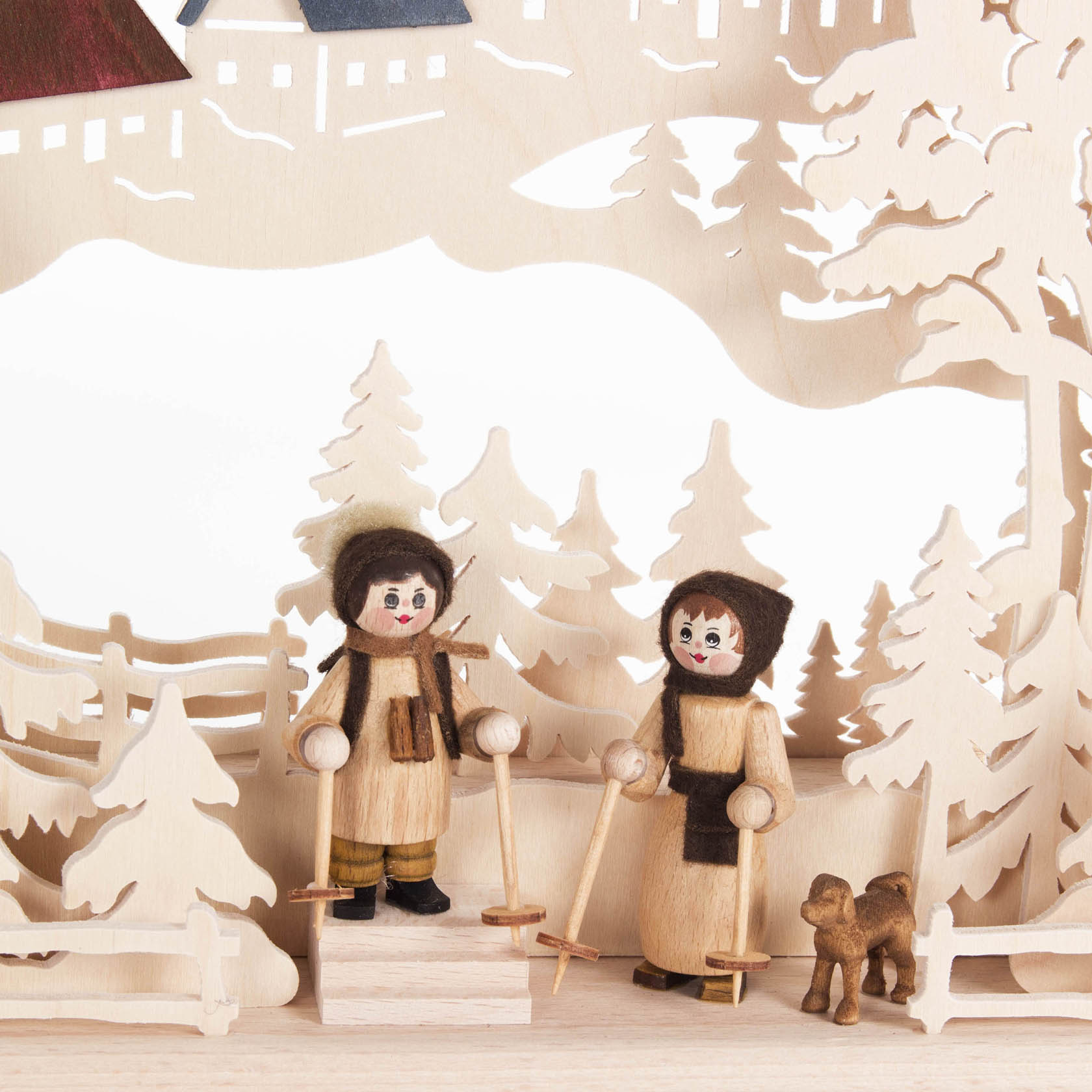 Two wooden figurines representing a man and a woman dressed for winter holding sleds, with two small dogs nearby, surrounded by a forest scene.
