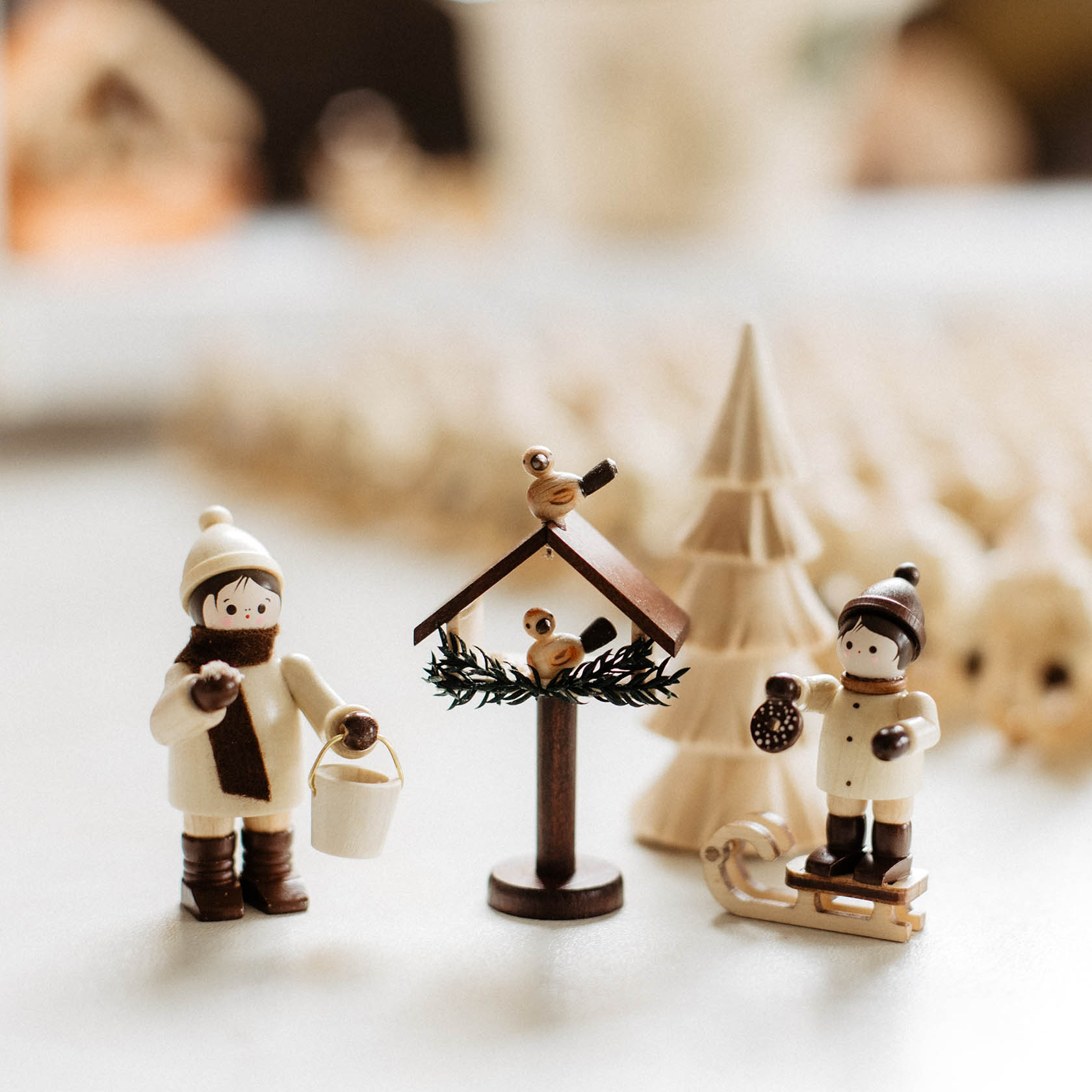 Holzfiguren in Winterkleidung, eine hält einen Eimer, die andere einen Schlitten, neben einem Vogelhaus mit einem Vogel oben und einem Baum.